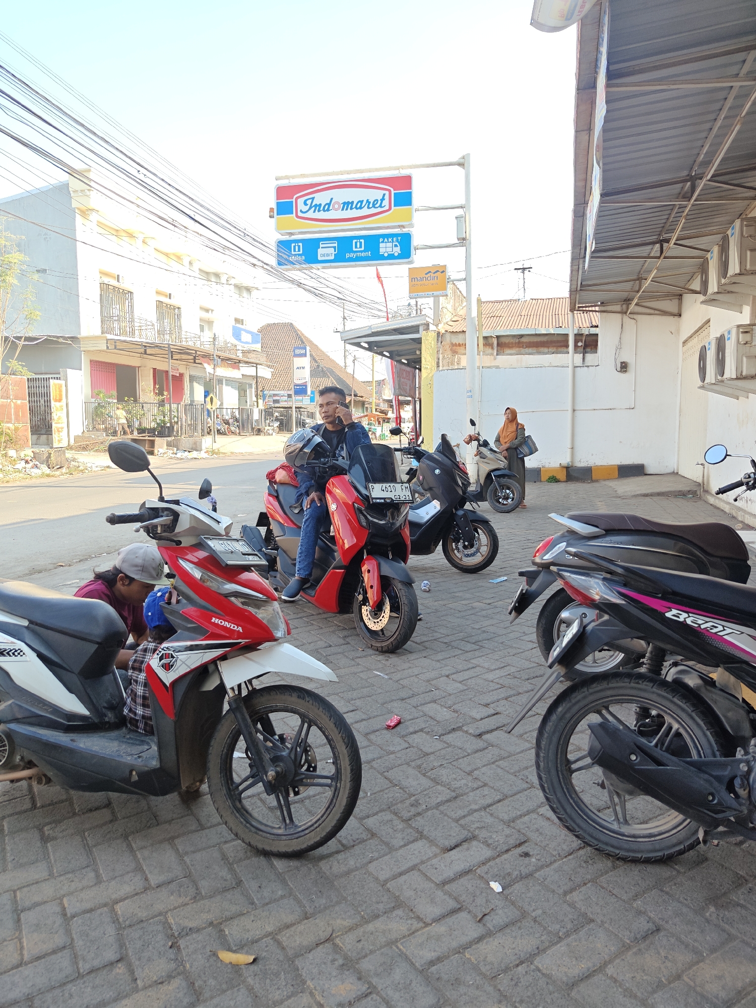 The image depicts a street scene in Klampokan, Indonesia, likely in front of an Indomaret convenience store. Several motorcycles are parked on a paved sidewalk. In the foreground, a red and white Honda scooter is partially visible, with a person in a grey cap hunched over its front wheel, appearing to be working on it. To the right, a man in a dark jacket and jeans is sitting on a red scooter, talking on a mobile phone. Behind him, another person in a headscarf stands near a beige scooter. In the background, buildings line the street, with power lines crisscrossing the sky. The overall impression is of a typical Indonesian street scene, with commerce and daily life unfolding.
