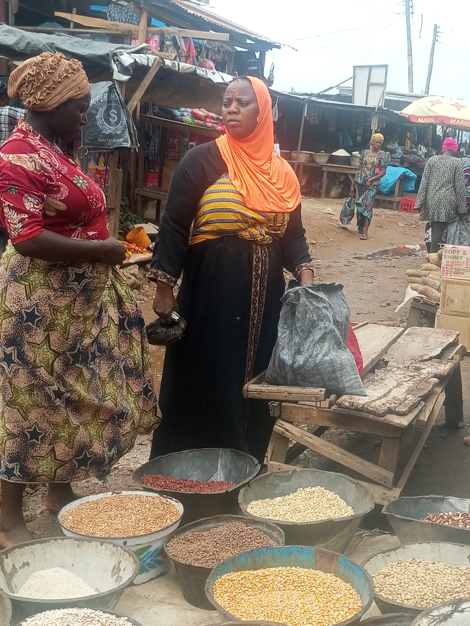In an outdoor market scene in Sabon Tasha, Nigeria, two women are the central figures. One woman is wearing a red floral print top, a patterned skirt, and a brown head wrap. She is standing to the left, looking down, with a small black object in her hand. The other woman is wearing a black dress with yellow striped detailing on the chest and sleeves, an orange head wrap, and holding a small black purse. She is looking to the right.
In front of the women are several metal bowls filled with various types of grains and beans. From left to right, there are bowls of white powder, light brown beans, dark brown beans, red beans, yellow beans, and white beans.
Behind the woman in black is a weathered wooden table with a gray sack on top. In the background, there are stalls with corrugated metal roofs, other people walking around, and a Maggi-branded umbrella. The ground appears to be dirt or gravel. A sign on a cardboard box reads "SOFT & SHINY."