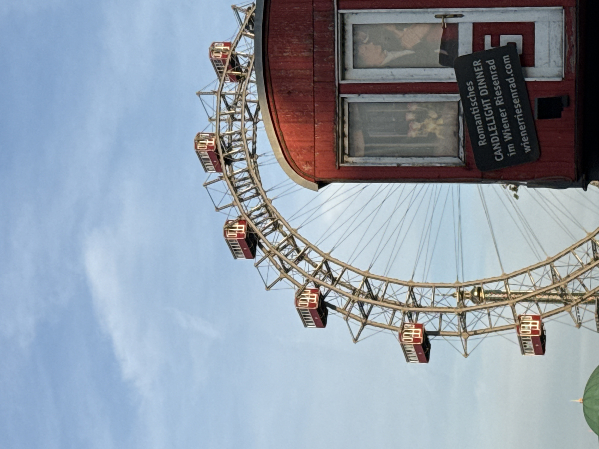 The image shows a segment of the Wiener Riesenrad, a large Ferris wheel located in the Prater amusement park in Vienna, Austria. The Ferris wheel is predominantly made of metal with red gondolas. In the foreground, a red carriage, likely part of the same attraction, is visible. This carriage has two windows, one showing a display with yellow flowers and another with a silhouette of a man and a woman. A black sign is attached to the carriage, which reads in white text: "Romantisches CANDLELIGHT DINNER im Wiener Riesenrad wienerriesenrad.com". The sky is a pale blue with scattered white clouds.