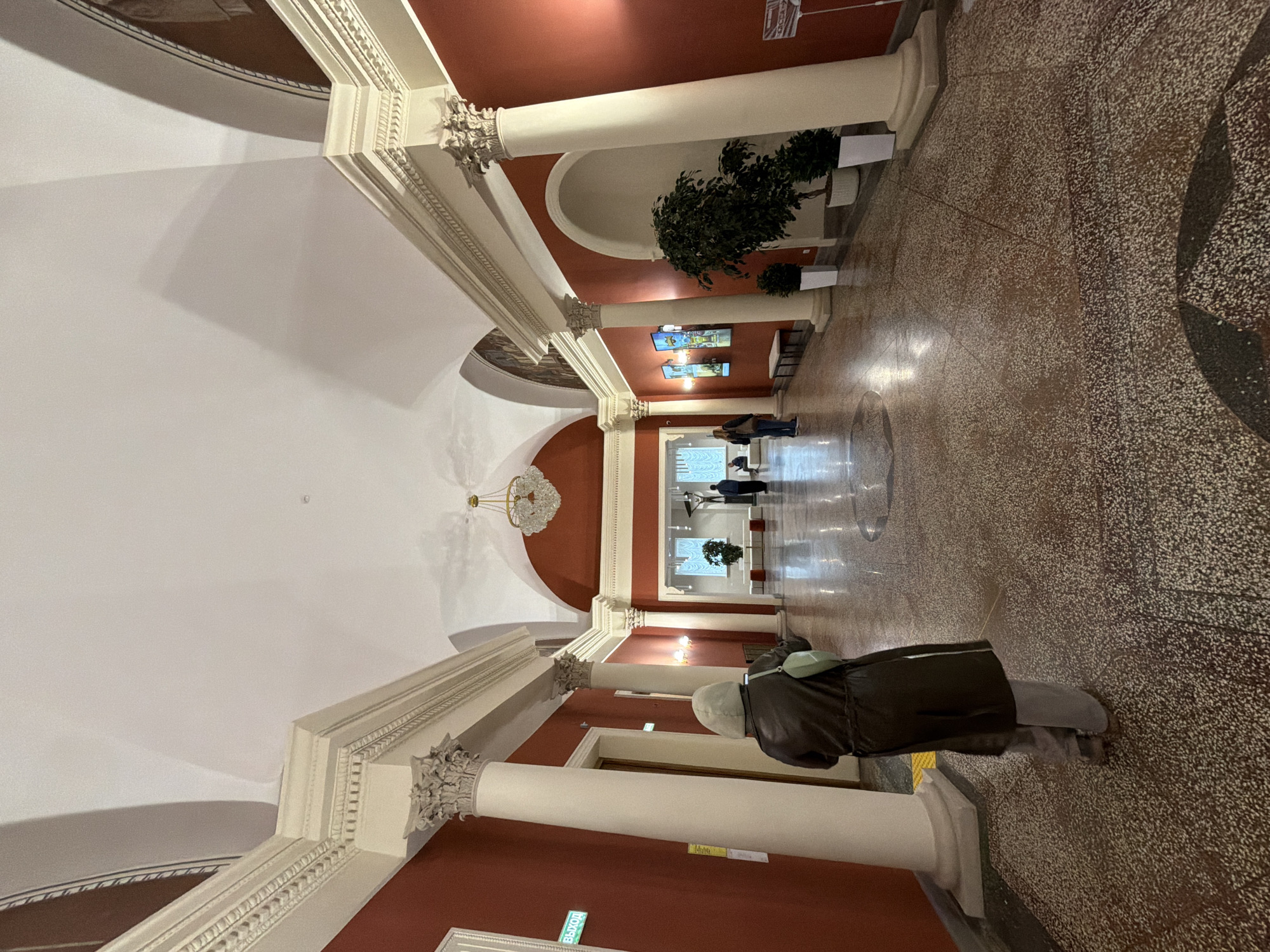The image shows the interior of a building featuring classical architecture, possibly a public space. It is marked by tall columns, arched doorways, and decorative molding along the ceiling. The walls are painted a deep red color above a white base, with the ceiling being white. The floor is a speckled terrazzo or similar material.
Several people are present, mainly in the distance, walking through the hallway or standing in front of what appears to be a display area. In the foreground, a person wearing a dark coat, light-colored pants, and a head covering is standing with their back to the viewer.
Potted plants are placed along the hallway, and video displays are mounted on the wall. The architecture and decor suggest that the location might be a museum, a government building, or a similar public institution. The city and exact location within the city are unknown.
