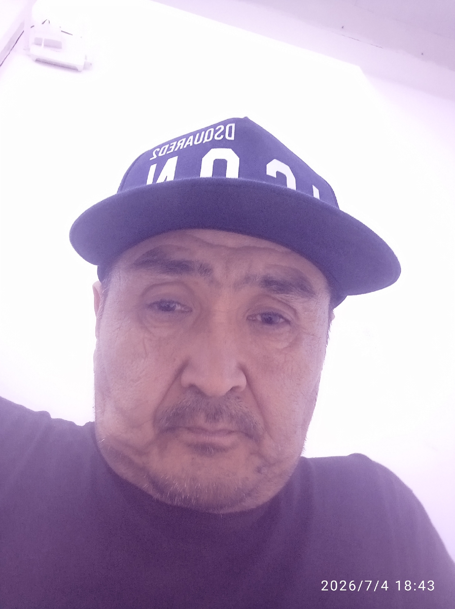 A close-up, waist-up, outdoor selfie shows a middle-aged man wearing a dark blue baseball cap and a dark purple t-shirt. The man has fair skin, dark hair, and a mustache. He is looking directly at the camera with a neutral expression. The background is a white wall. The date and time stamp on the bottom right corner of the image reads "2026/7/4 18:43".