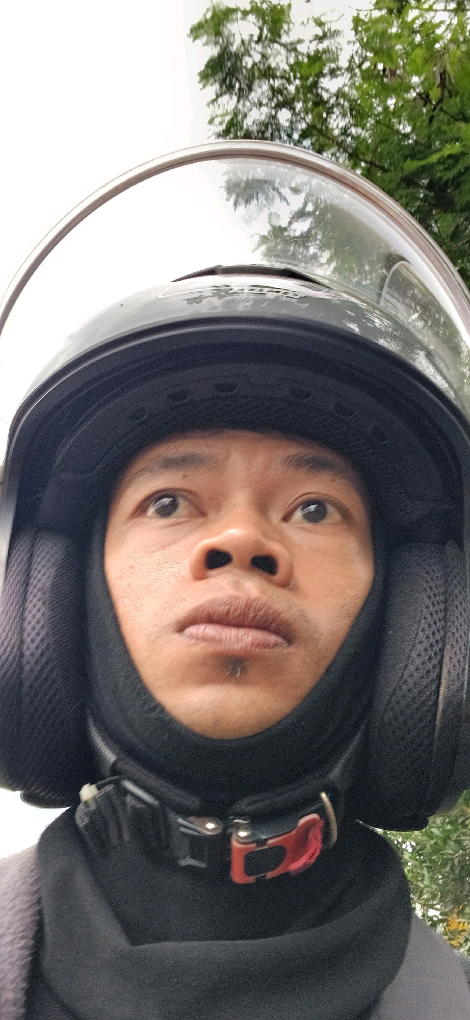 A close-up, slightly upward-facing selfie shows a person wearing a black motorcycle helmet with a clear visor. The person's face, with dark eyes and visible stubble on their chin, is framed by the helmet and a black fabric covering that extends up to their nose. The helmet has black ear pads, and a red and black buckle is visible at the neck. In the background, out-of-focus green trees and a bright, overcast sky are visible, suggesting an outdoor setting, potentially in a rural area given the foliage. The context implies the person is likely riding a motorcycle. The image has a resolution of 2268x4032 pixels and was taken on an unknown date, possibly in or around Wonokerso, Indonesia.