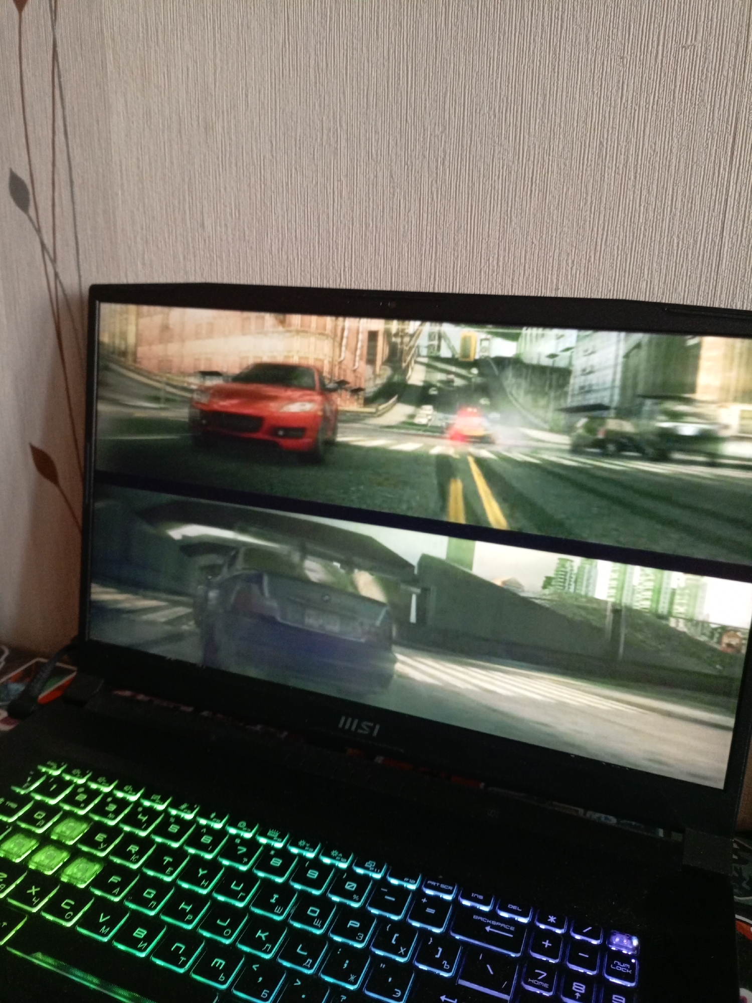 Here's a detailed description of the image:
The image shows a laptop with a gaming keyboard featuring multicolored backlighting. The screen displays two segments of a racing game, possibly Need for Speed.
* **Top Segment:** A red sports car is positioned on a city street with other vehicles in the distance.
* **Bottom Segment:** A dark-colored car is seen from the rear, also on a city street.
The laptop is resting on a surface with indistinct objects underneath. The background is a neutral-colored wall with a decorative plant design. The brand MSI is visible on the laptop. The laptop keyboard is Russian layout.
There is no indication of the image being taken in the city of Saratov, Russia.
