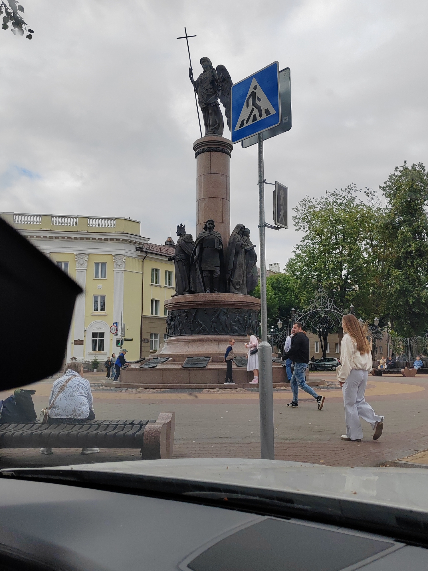 The image depicts a street scene in Brest, Belarus, likely near the monument to the Millennium of Brest, which features an angel holding a cross at its apex. In the foreground, a person is seated on a bench, their back to the camera, wearing a light-colored jacket with a patterned design. To the left, a large black object, possibly an umbrella, partially obscures the view.
A pedestrian crossing sign, a blue square with a white depiction of a person walking, is prominently displayed on a pole. Several people are visible walking in the middle ground. A group of individuals, including two women and a man, are walking near the base of the monument. Another man in jeans and a dark jacket is walking past the pedestrian sign. A woman in light-colored pants and a white top walks towards the right side of the frame.
The monument itself is a large, multi-tiered structure with several sculpted figures at its base, including a king and other historical figures. Behind the monument, a light yellow building with white architectural details and numerous windows is visible. Trees with green foliage line the background, and the sky is overcast. The ground is paved with reddish-brown bricks. The scene suggests a public square or thoroughfare in an urban environment.