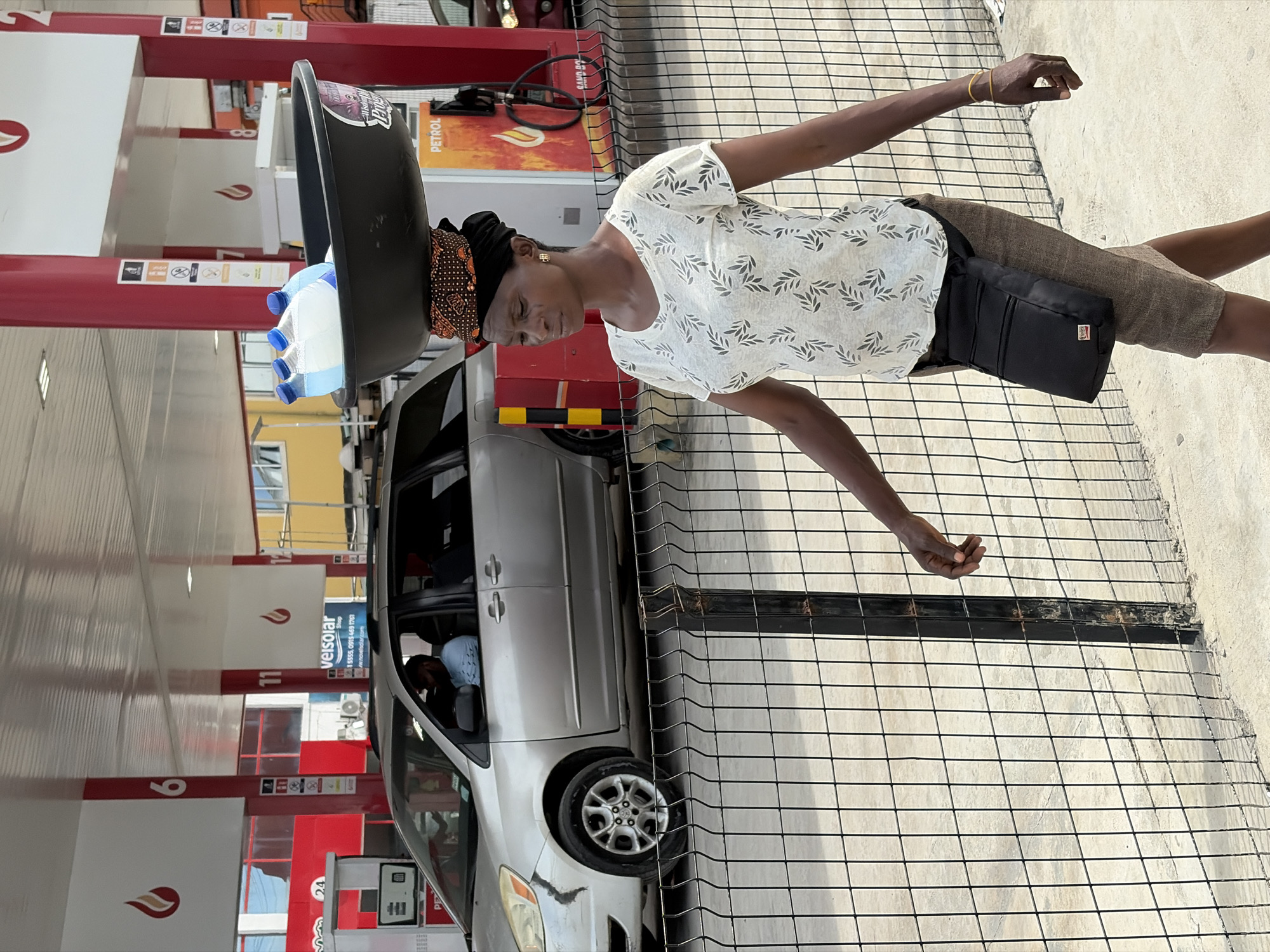 In Igboefon, Nigeria, a woman carries a large black basin filled with blue and white plastic containers on her head at a Conoil petrol station. She wears a light-colored patterned shirt, khaki shorts, and a black bag around her waist. She stands near a silver minivan and a black metal fence. The petrol station has red and white pump stations.