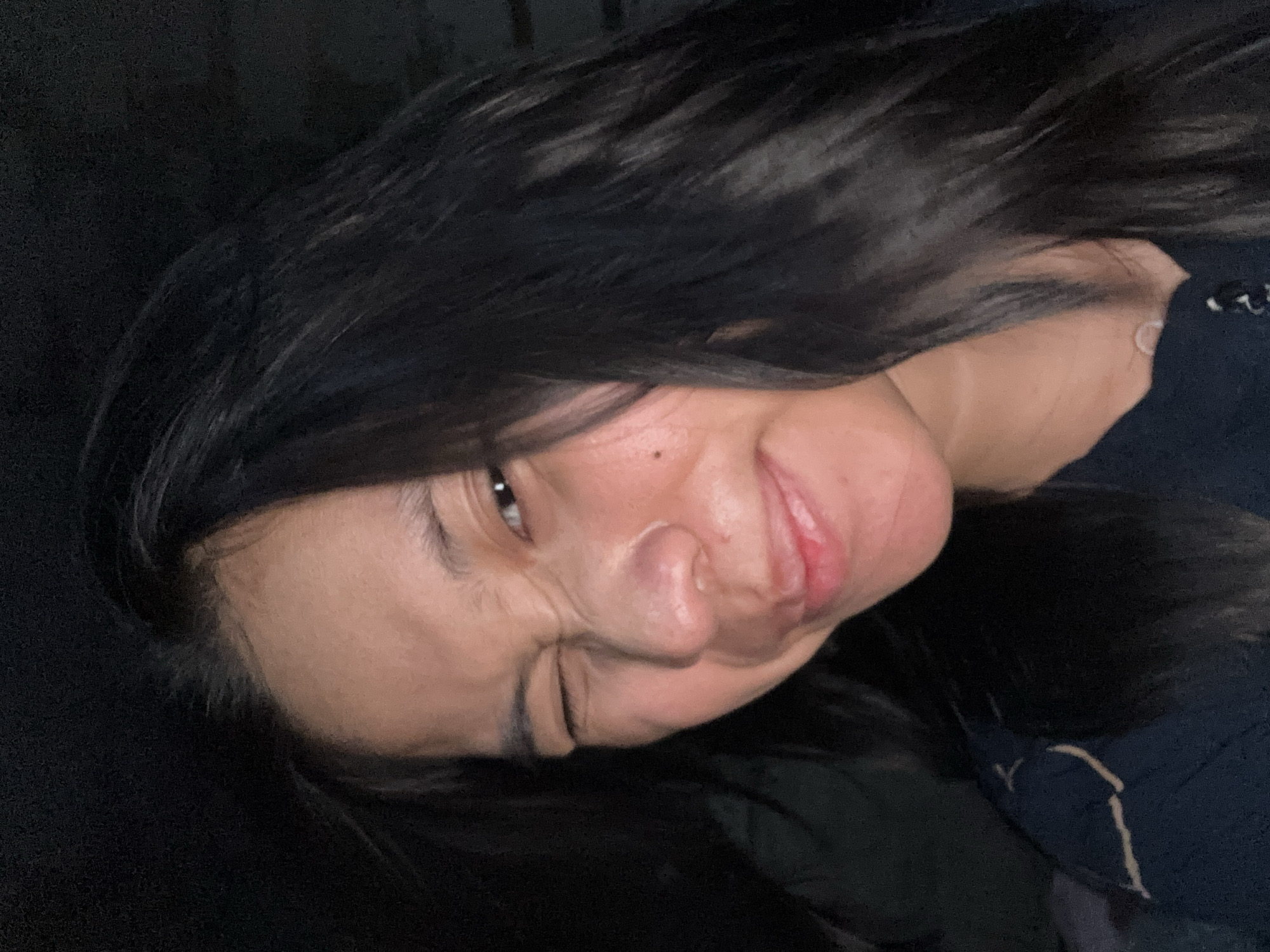 The image is a close-up selfie of a young East Asian woman with long, dark hair. She is winking and smiling, with her nose slightly scrunched. Her skin appears to have some texture and a few small blemishes. She is wearing a dark-colored top with some lighter colored trim on the shoulder area. The background is dark and out of focus, suggesting the photo was taken in low light conditions. There are no identifiable elements in the background that would indicate a specific location in East Jakarta, Indonesia.
