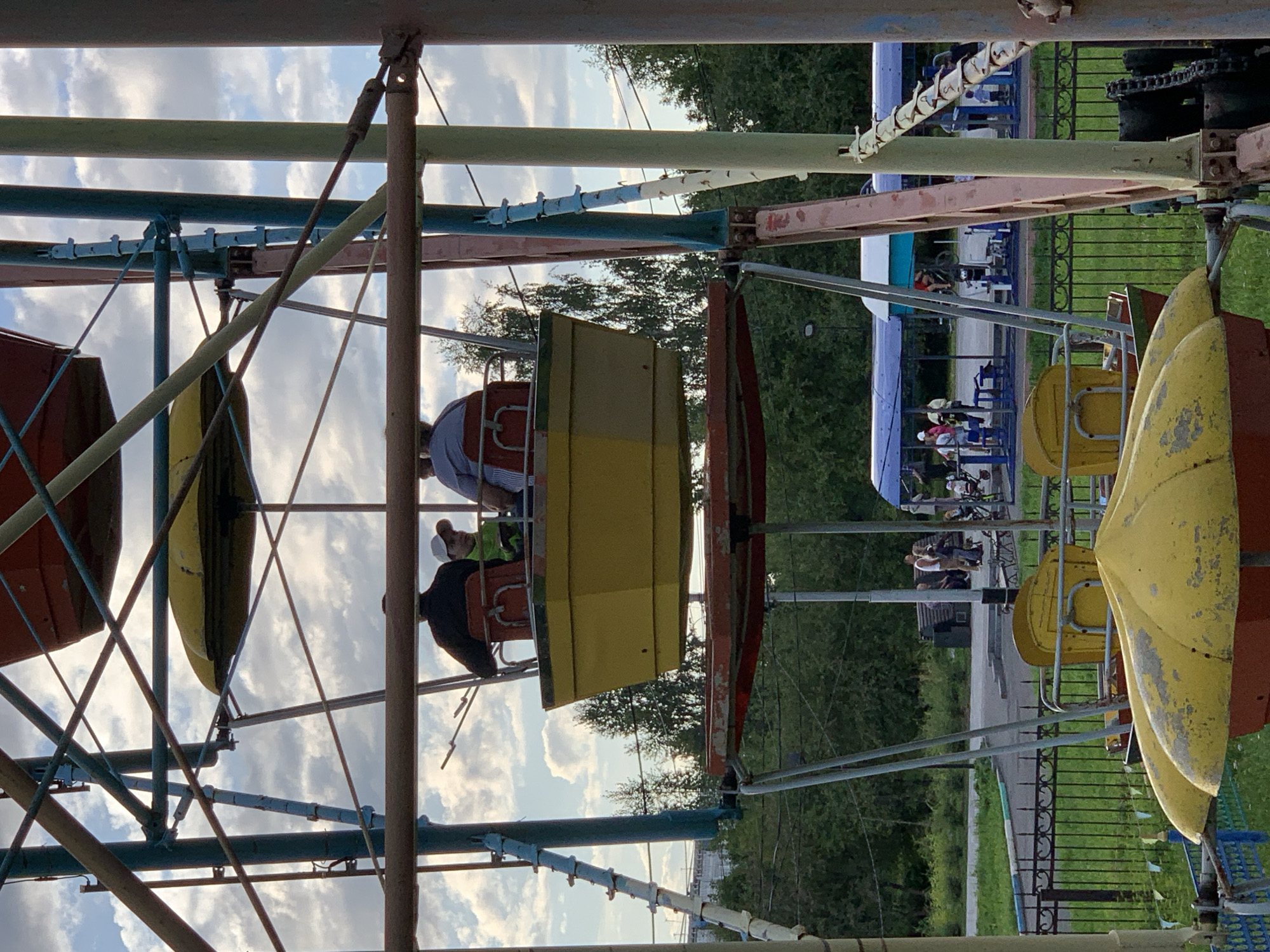 This image depicts a Ferris wheel ride at the amusement park in Prokopyevsk, Russia. The Ferris wheel has a weathered, colorful appearance, with yellow and red gondolas. In the foreground, two yellow, bell-shaped structures, possibly part of a different ride, are visible, partially obscuring the lower portion of the Ferris wheel.
In the middle section of the Ferris wheel, a yellow gondola is carrying three individuals. The person in the front is wearing a black jacket, the person behind them is wearing a green shirt and a white cap, and the person in the rear is wearing a light blue shirt. They are all seated and facing forward, appearing to be enjoying the ride.
In the background, a row of trees and a blue and white structure, possibly another amusement ride or shelter, are visible. Several other people are seen in the background, some standing and observing, while others appear to be walking. A black metal fence runs across the middle ground, separating the amusement park area from a grassy lawn. The sky is partly cloudy, with patches of blue sky visible. The overall impression is of a functioning, albeit somewhat dated, amusement park.