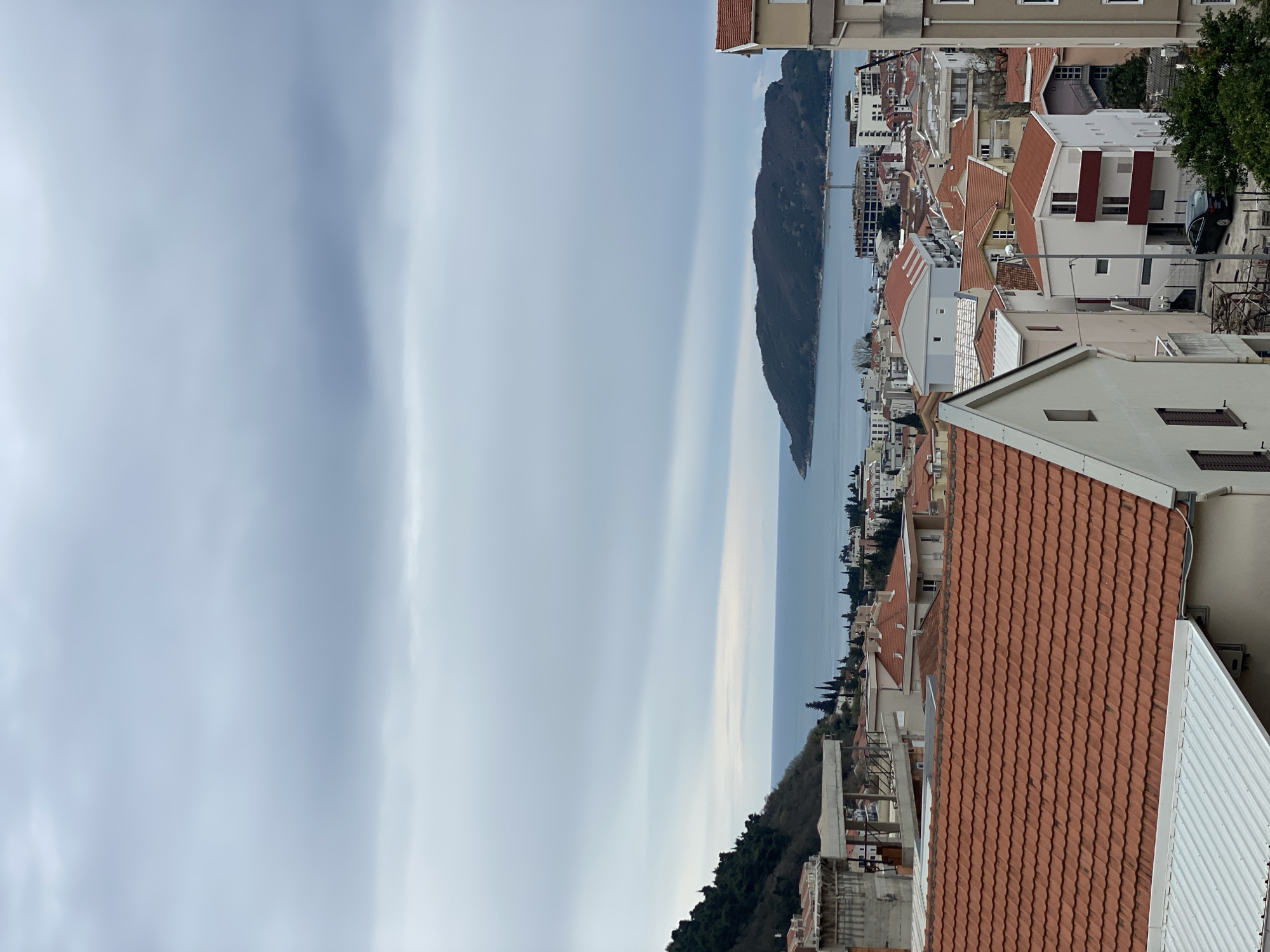 The image depicts an aerial view of Budva, Montenegro, focusing on a cityscape with a mountain and sea in the background. The sky is overcast with prominent cloud formations. In the foreground, there are buildings with orange and white roofs. The buildings are densely packed together, indicative of a coastal urban area. The mountain, covered with vegetation, extends into the sea. A series of buildings appears on the other side of the mountain. The sea itself is a pale blue color, reflecting the overcast sky. There are no people visible in the image.