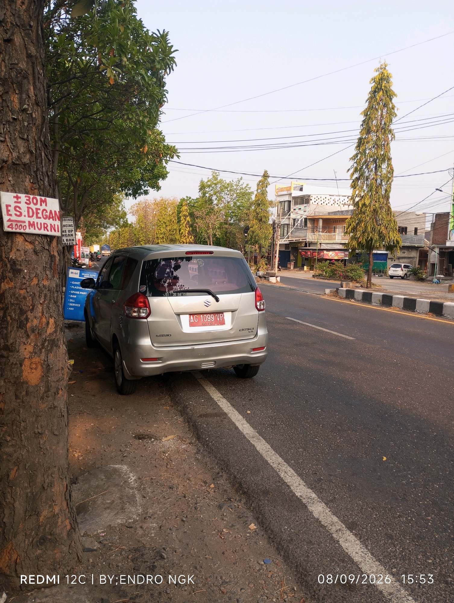 The image is a roadside scene in Ringinanom, Indonesia, captured at 3:53 PM on September 8, 2026. A silver Suzuki Ertiga is parked on the side of a paved road, angled slightly away from the viewer. The car's rear is visible, displaying a license plate that reads "AG 1099 VP". To the left of the car, a large tree trunk with rough bark is prominent, with a red and white sign attached to it. This sign has some text, including "30M ES:DEGAN" and "4000 MURNI". Below the first sign, a blue sign with white text is partially visible, listing services like "MOBIL", "FLASHING", "SPARE PART", "BAN", "DLL", and "SERVICE". The road has a white dashed line marking the lane and a black and white concrete barrier along its edge. In the background, there are buildings with multiple stories and trees, including tall, slender trees with yellow-green foliage. Power lines crisscross the sky. The ground next to the road is unpaved, with dirt and small stones. The lighting suggests it is daytime, with sunlight casting shadows. The bottom right of the image includes the text "08/09/2026 15:53" and "REDMI 12C | BY:ENDRO NGK".