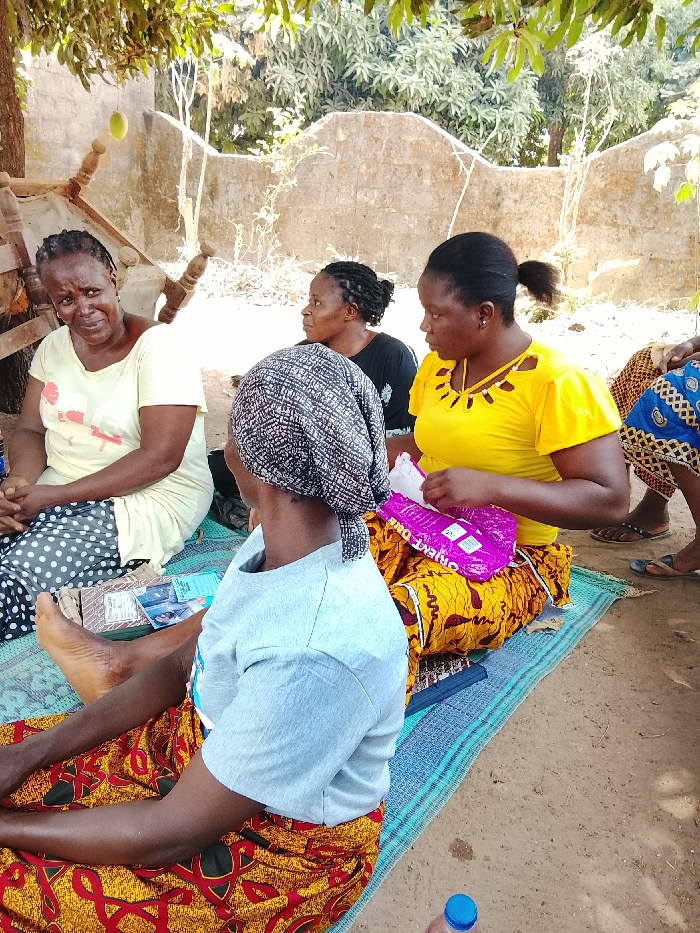 An outdoor scene in Akwanga, Nigeria, depicts five adult females seated on blue and green striped woven mats on a dirt ground under a tree.
On the far left, an adult female with braided hair, wearing a light yellow short-sleeved top and a black and white polka dot skirt, looks towards the viewer with a neutral expression, her hands clasped. To her right, another adult female, partially obscured and in profile, wears a black top and observes the scene to her right. Behind her, a dilapidated wooden frame is visible under a tree branch from which a green mango hangs.
In the foreground, an adult female with her back to the viewer wears a patterned headscarf, a light grey short-sleeved t-shirt, and a red and yellow patterned wrapper skirt; she is barefoot. To her right, an adult female in a yellow short-sleeved top with cut-out neckline details and a yellow and orange patterned wrapper skirt is actively handling a purple plastic bag, revealing the partial text "ORIENT ONE." Her hair is tied back. To the far right, only the legs and lower portion of a blue patterned wrapper skirt are visible, with dark sandals on the feet.
On the mat between the leftmost woman and the woman with her back to the viewer, several folded Nigerian Naira currency notes and a stack of pamphlets or documents are arranged. A blue-capped plastic water bottle rests on the dirt ground in the bottom right corner. The background features a low, weathered concrete wall and dense green foliage under natural daylight conditions.