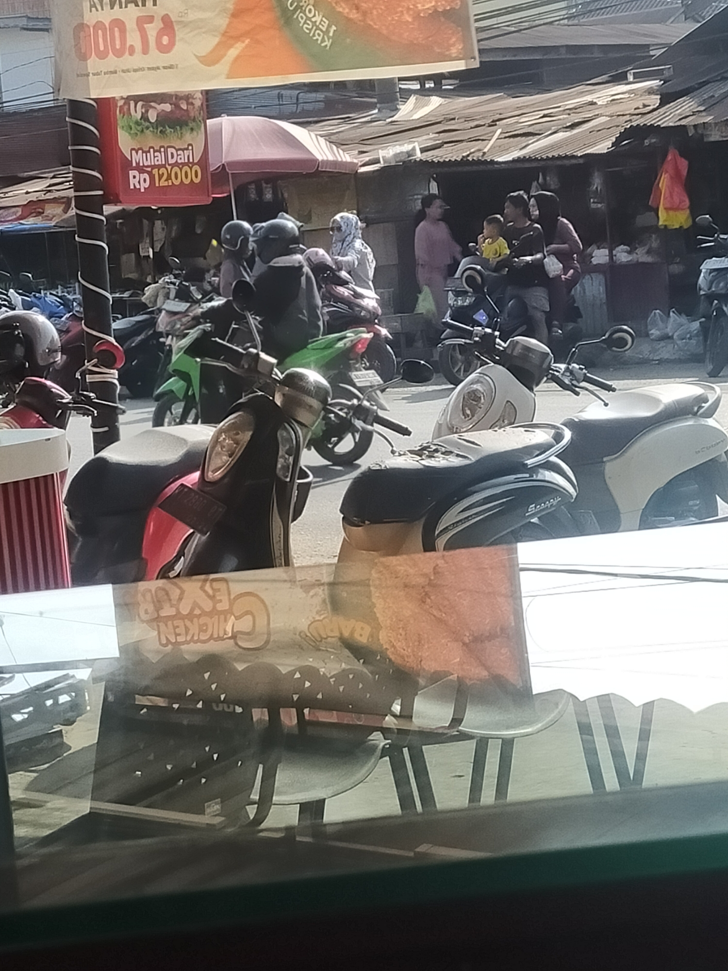The image depicts an outdoor scene in what appears to be a marketplace or a street in an urban setting, likely in Southeast Asia. In the foreground, a red and white striped cylindrical object is visible on the left. Behind it, several motorbikes are parked. One motorbike in the center is black and red, with a black seat. Further back, a green motorbike is parked.
In the middle ground, there is a row of stalls with corrugated metal roofs. People are gathered around these stalls. On the left, a person wearing a pink headscarf stands near a stall. To their right, a man and a child are on a motorbike. Another person, wearing a darker headscarf, is behind them. To the right of this group, there is another stall with items displayed.
In the background, more stalls and buildings are visible, along with a clear sky. A large banner with text and an image of a burger is hanging from a pole on the left. Another banner with text and an image is above the stalls. The overall impression is of a busy street scene with commerce and people on motorbikes. The lighting suggests it is daytime.