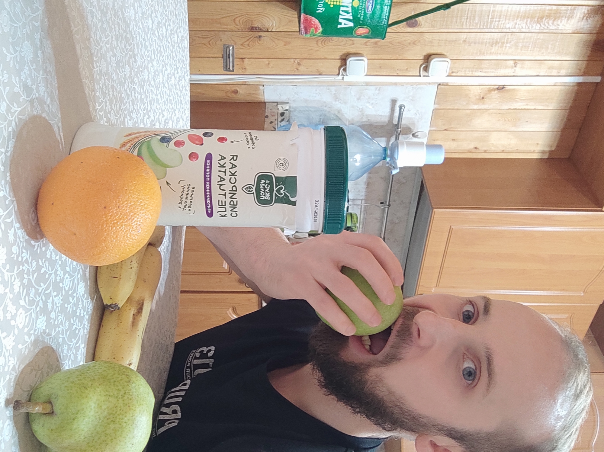 A man is biting into a green apple, while an orange, a cluster of bananas, a green pear, and a large bottle of liquid are on a table to the left. The table is covered with a patterned white tablecloth. The background features wooden walls and cabinets, along with a water dispenser setup. A green container with a strawberry illustration is visible in the upper part of the background.