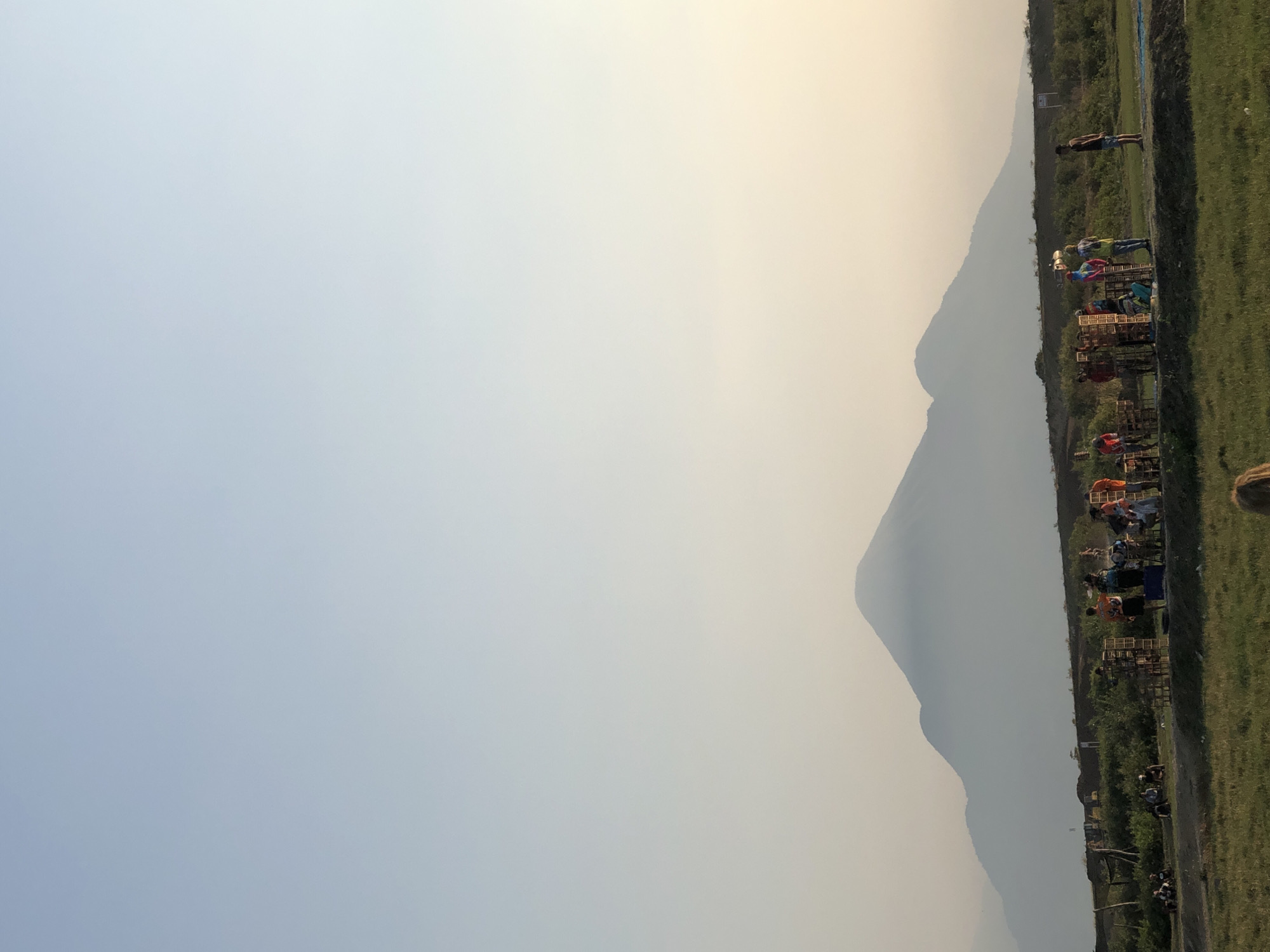 In Gempolsari, Indonesia, a group of people are gathered in a grassy field, preparing what appear to be fireworks. A large, mist-shrouded mountain range dominates the background. The sky is a pale blue, fading into a soft yellow and orange near the horizon, suggesting either dawn or dusk. Several individuals are actively involved in arranging numerous wooden crates, likely containing fireworks, on the ground. Others are standing or sitting nearby, observing the preparations. The environment is rural, with sparse vegetation and a faint haze in the air. One person stands apart, facing away from the group towards the landscape.