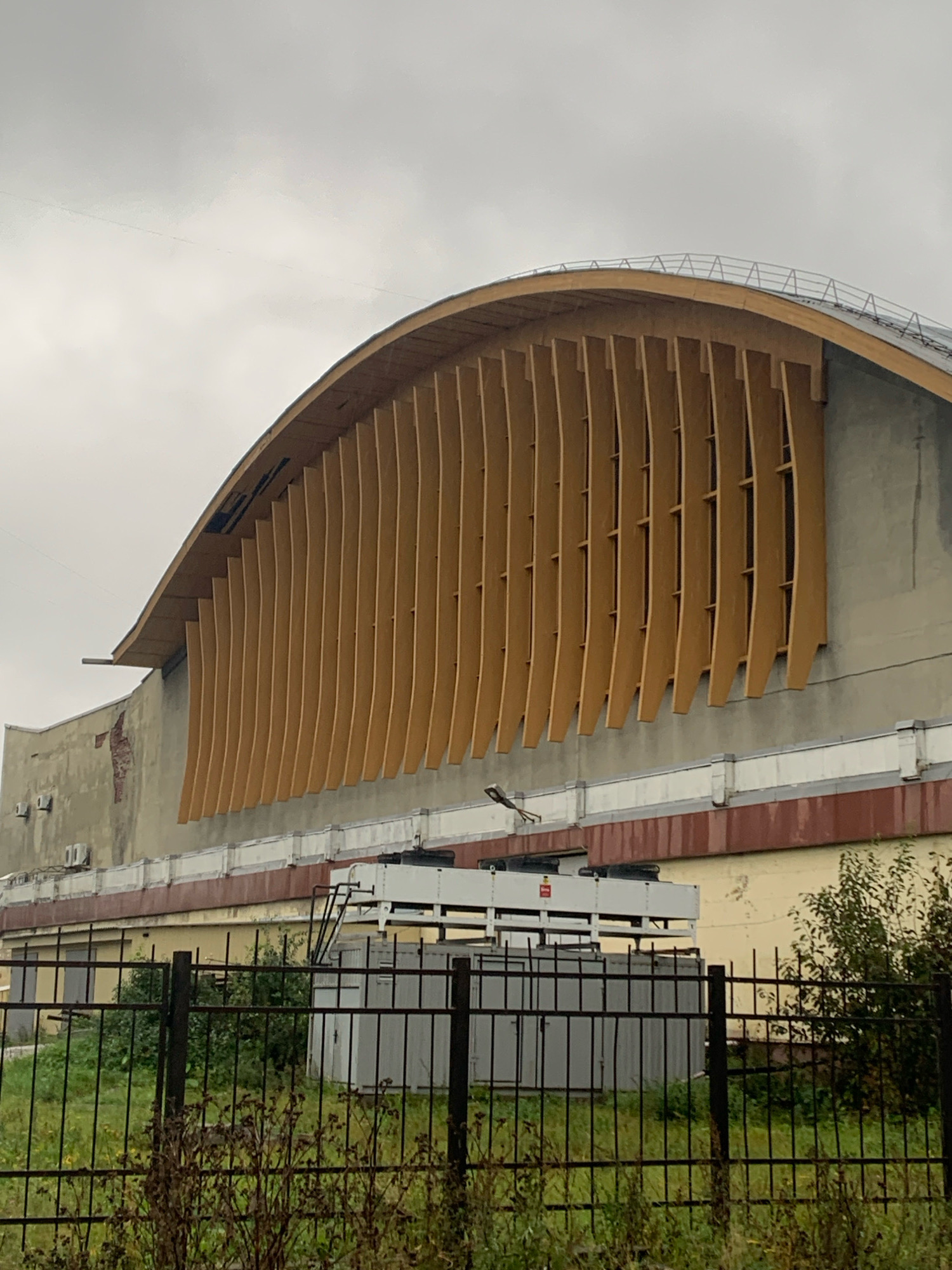 The image shows the side view of a large, arched building with a distinctive wooden slat facade. The building appears to be a sports or event venue. The wooden slats are vertically oriented and curved, creating a textured and visually appealing exterior. Below the wooden facade, the building has a more traditional concrete structure. There is a prominent white band running horizontally along the upper section of the concrete wall, and below that, a darker reddish-brown band. In front of the building, there is a black metal fence surrounding an area with overgrown grass and some dry bushes. Behind the fence, there are several industrial-looking structures, including large grey containers and white equipment that resembles air conditioning units or machinery. The sky is overcast and grey, suggesting a cloudy day. The overall impression is of an urban industrial or institutional setting. This building is the Arkhangelsk Ice Palace located in Arkhangelsk, Russia.