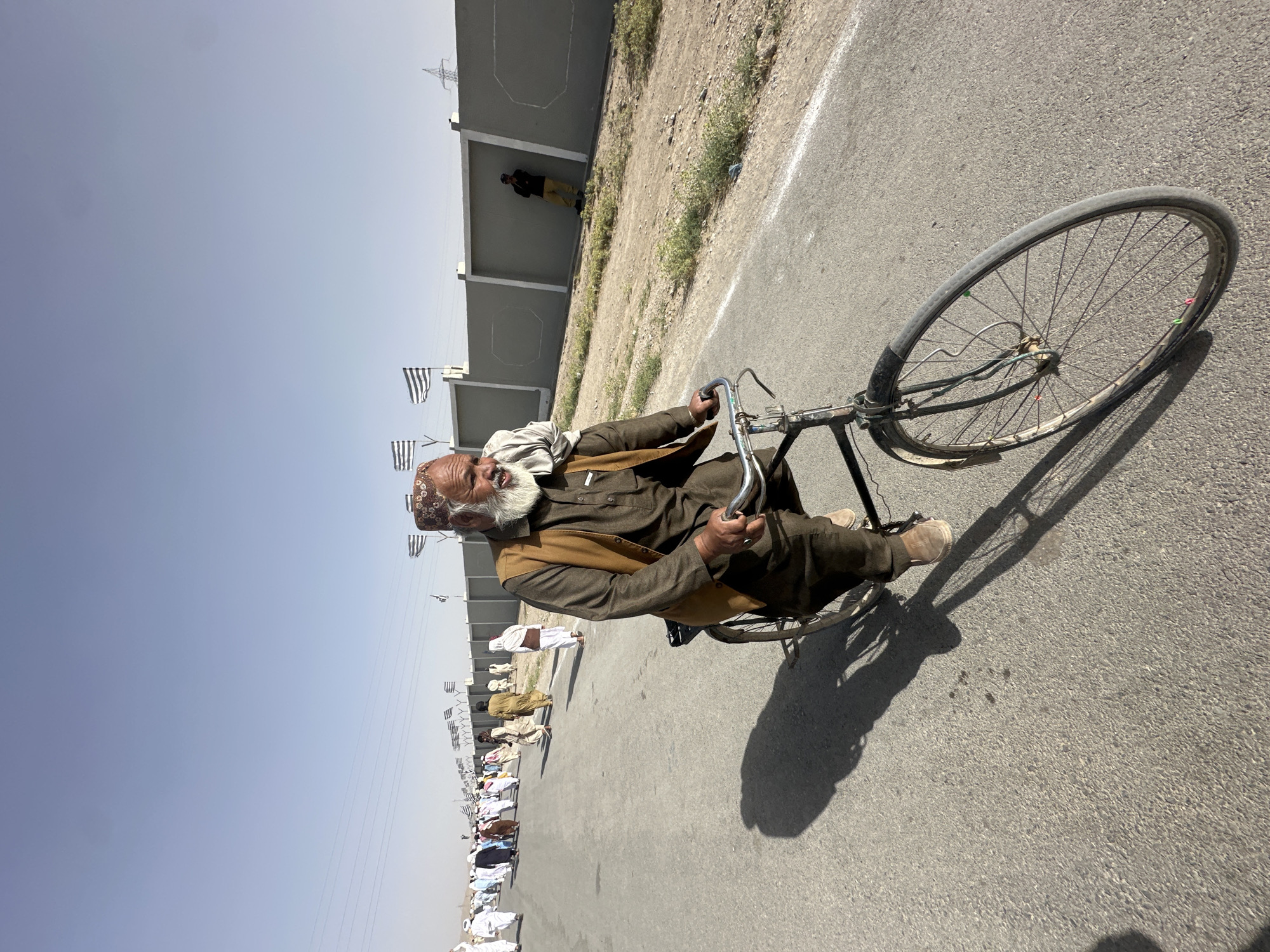 Captured in Pishin, Pakistan, the image depicts an elderly man riding an old bicycle on an asphalt road under a bright, clear sky. The man, positioned prominently in the foreground, has a long white beard, wears glasses, and is dressed in traditional attire including a dark brown kurta, a light brown waistcoat, and a patterned cap. He is actively pedaling, gripping the handlebars of his simple, well-used bicycle.
Strong shadows are cast on the road, indicating bright sunshine and suggesting it is likely mid-day. Along the left side of the road, a line of other individuals, also in traditional clothing, can be seen in the background, appearing to walk or gather. A low concrete wall runs parallel to the road, adorned with several small, black and white striped flags attached to poles. Beyond the wall lies dry, uncultivated ground, with power lines visible in the distant sky. Another person stands near the wall in the mid-ground. The overall scene captures a moment of daily life, with no specific event immediately apparent beyond people moving along the road. No visible text is present in the image.