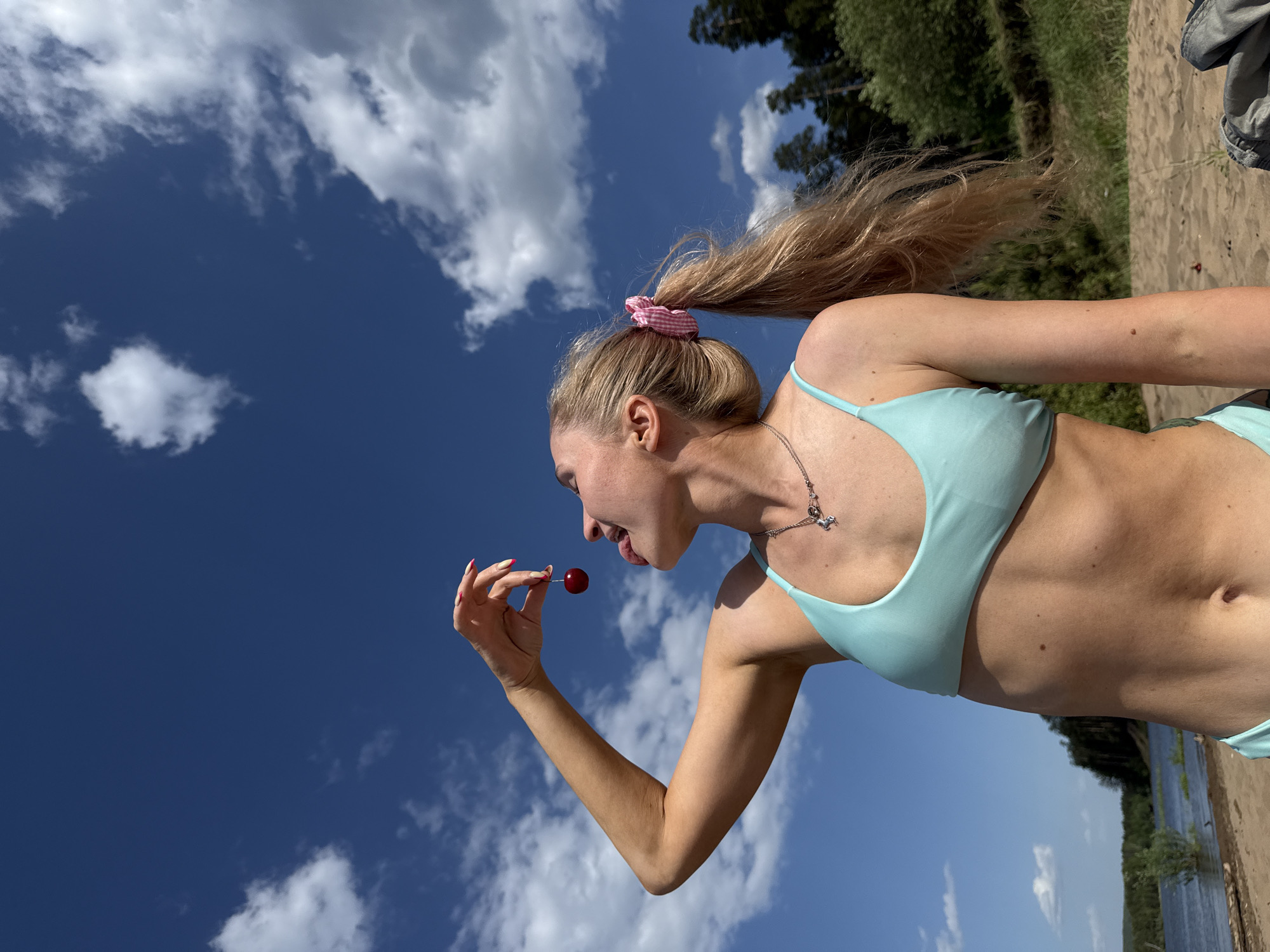 A slender woman with long blonde hair pulled into a high ponytail held with a pink scrunchie is positioned against a background of bright blue sky dotted with fluffy white clouds. She is wearing a light blue bikini. She is holding a red cherry in her left hand and about to put it in her mouth. She has a silver necklace with a small pendant. The photo appears to have been taken outdoors in Chaikovsky, Russia, near a body of water with a sandy bank and green trees along the shore.