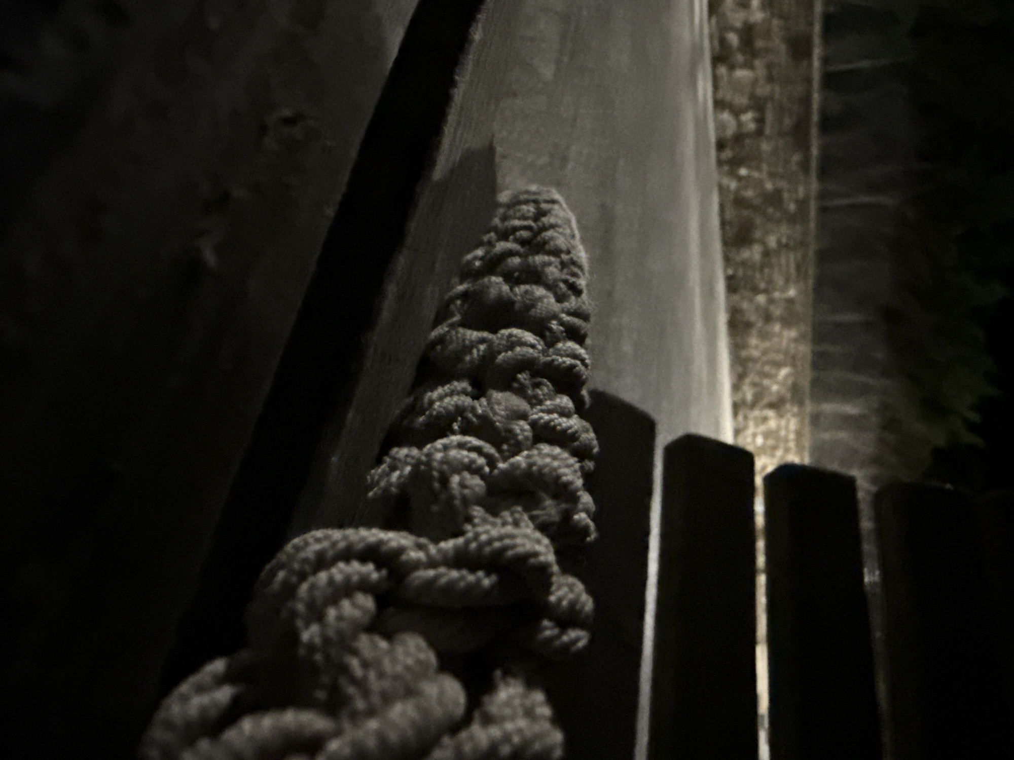 An outdoor night scene in Tyulenovo, Bulgaria, shows a thick, braided rope lying horizontally across a dark wooden surface. The rope is textured and appears to be made of natural fibers. To the left of the rope, a paved surface is visible, with a low stone wall and trees in the background, illuminated by ambient light. To the right of the rope, the dark, slatted structure of a bench or seating area is present. The lighting is dim, with shadows obscuring details in the foreground and background, creating a moody and somewhat abstract composition. No people are present in the image.