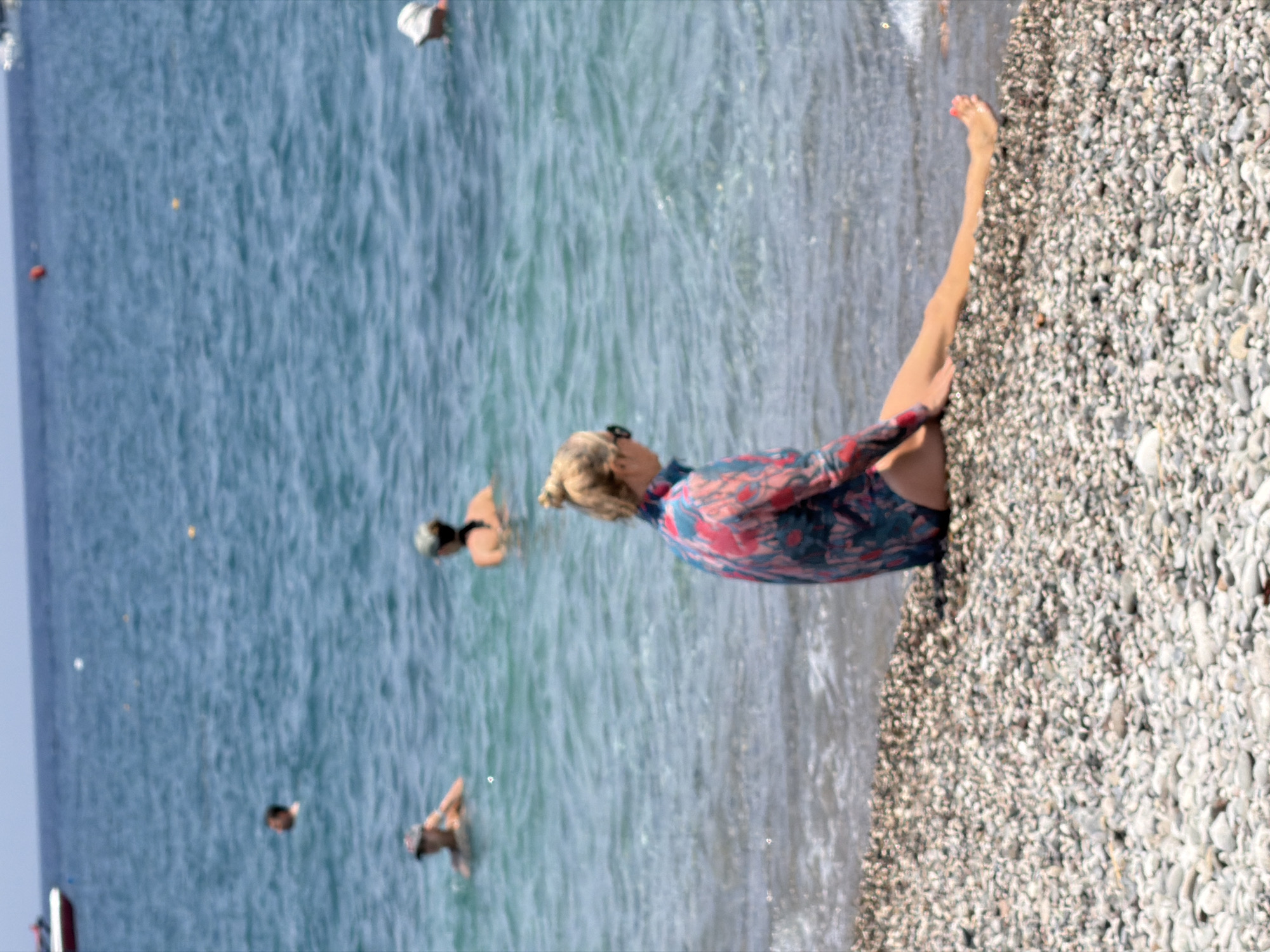 A woman with blonde hair pulled into a bun, wearing sunglasses and a colorful rash guard, sits on a pebble beach in Kemer, Turkey, facing the sea. She is positioned with her legs extended towards the water, with her right foot slightly submerged. Several other people are visible in the turquoise water, some swimming and others appearing to be standing. A white boat is docked in the distance on the left. The sky is clear and blue, indicating a sunny day.