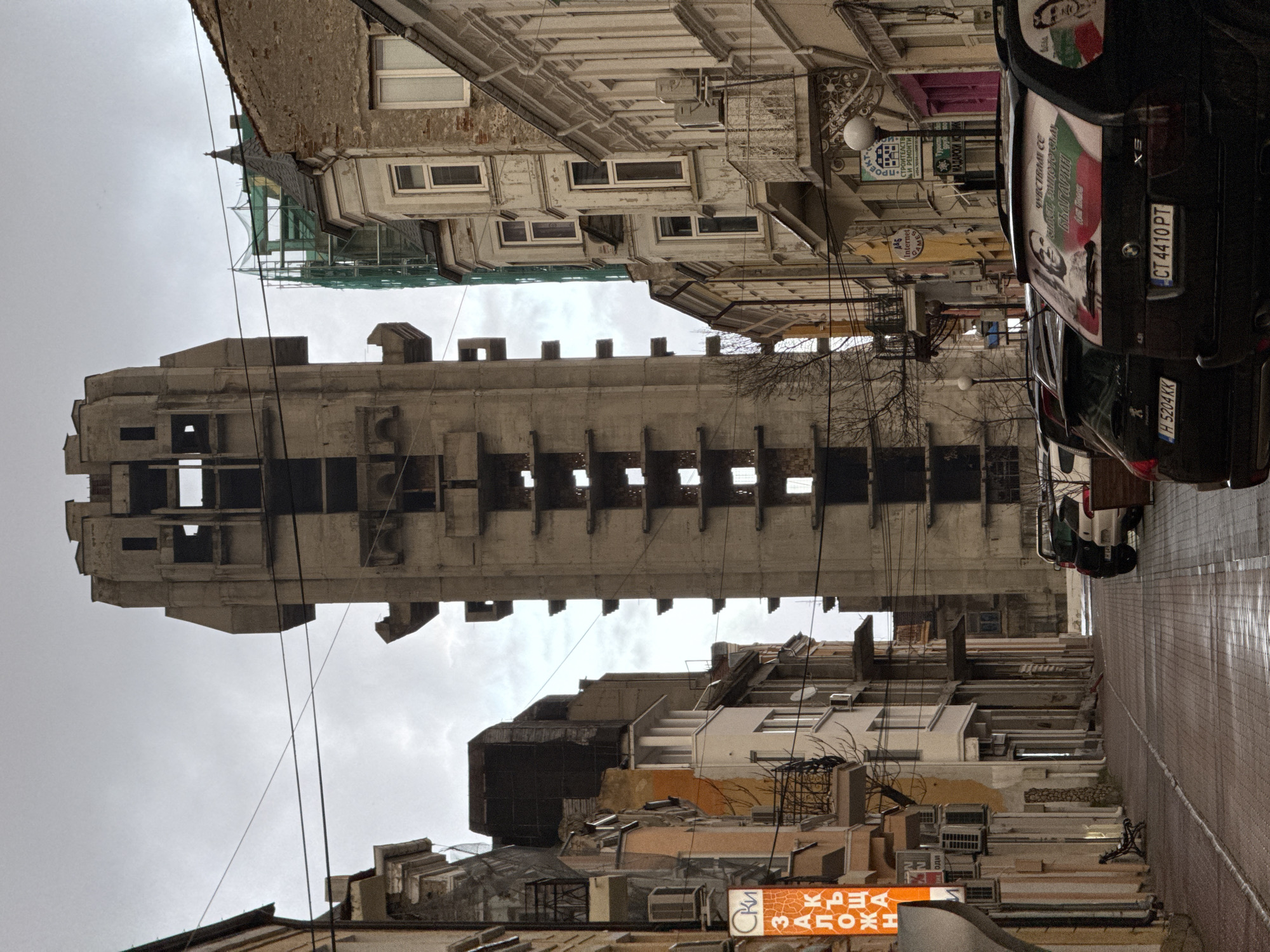 The image shows an urban street scene in Shumen, Bulgaria, dominated by a large, unfinished concrete building in the center, its bare structure rising above the surrounding buildings. The street is lined with buildings of varying heights and architectural styles, some appearing older and in need of repair. There are multiple overhead wires crossing the street, adding to the visual clutter. Several parked cars are visible on the right side of the street, including a black BMW with stickers related to Bulgaria. The sky is overcast and gray. Signage in Bulgarian is visible, adding context to the location. The street appears wet, possibly from recent rain.