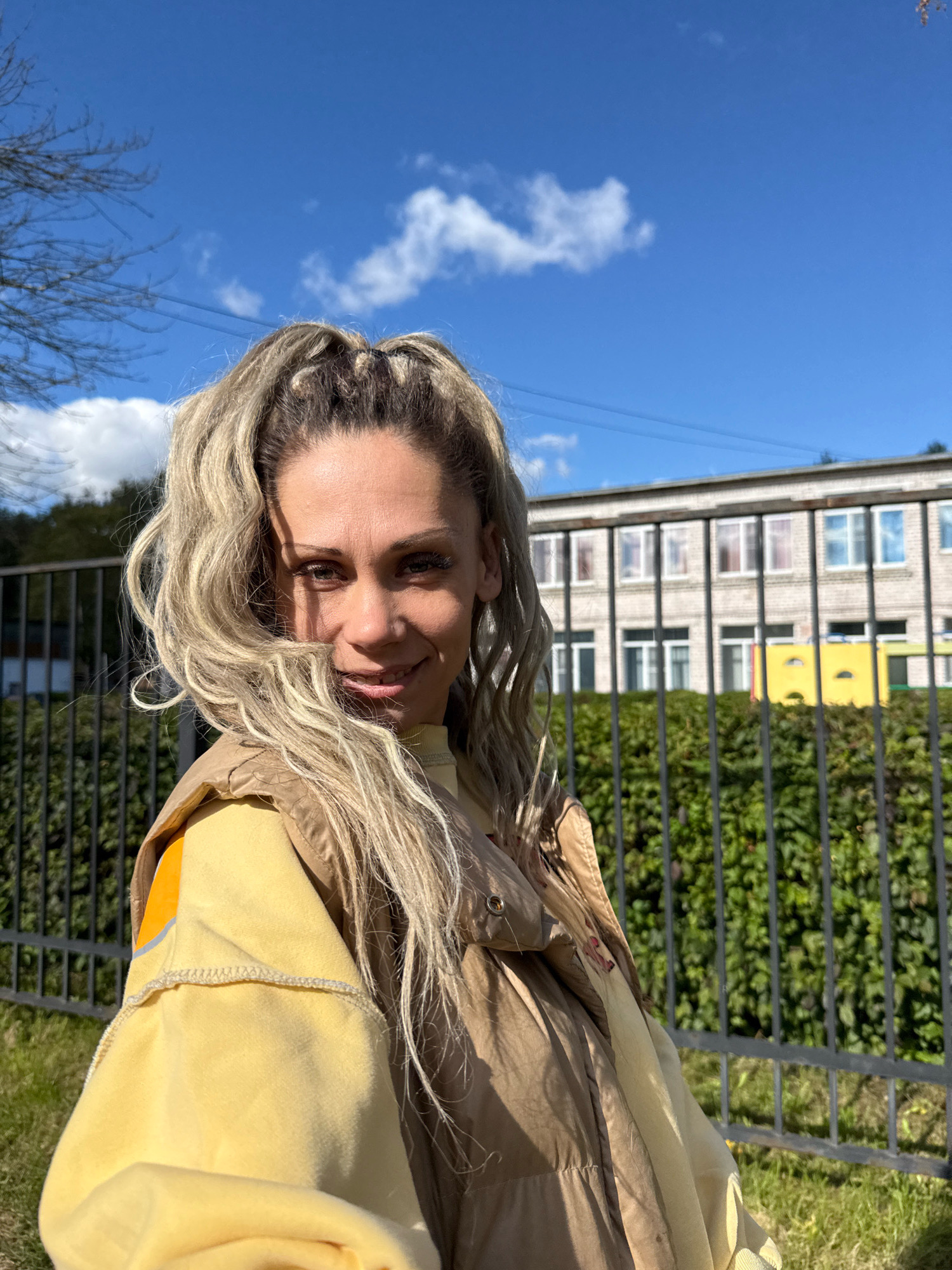 A woman with blonde, wavy hair pulled into a high ponytail is taking a selfie outdoors. She is wearing a yellow sweatshirt and a brown puffer vest. She is smiling and looking at the camera. Behind her, there is a black metal fence, a green hedge, a building with many windows, and a blue sky with white clouds. The scene appears to be located in Rusanovo, Russia.
