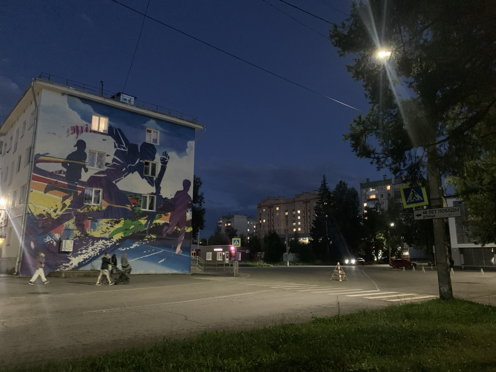 The image depicts a street scene in Asha, Russia, at dusk. A large building with a colorful mural on its side dominates the left portion of the frame. The mural features silhouetted figures and abstract shapes in vibrant colors, with some windows of the building illuminated. Several people are visible walking on the sidewalk and across a street.

The street is paved and appears to be an intersection, with crosswalks marked. Traffic lights and street signs are present. In the background, apartment buildings with illuminated windows are visible, suggesting a residential area. Trees line the street, and a streetlamp illuminates the scene with its bright light. A car is seen driving on the street. The overall atmosphere is that of a quiet urban evening.
