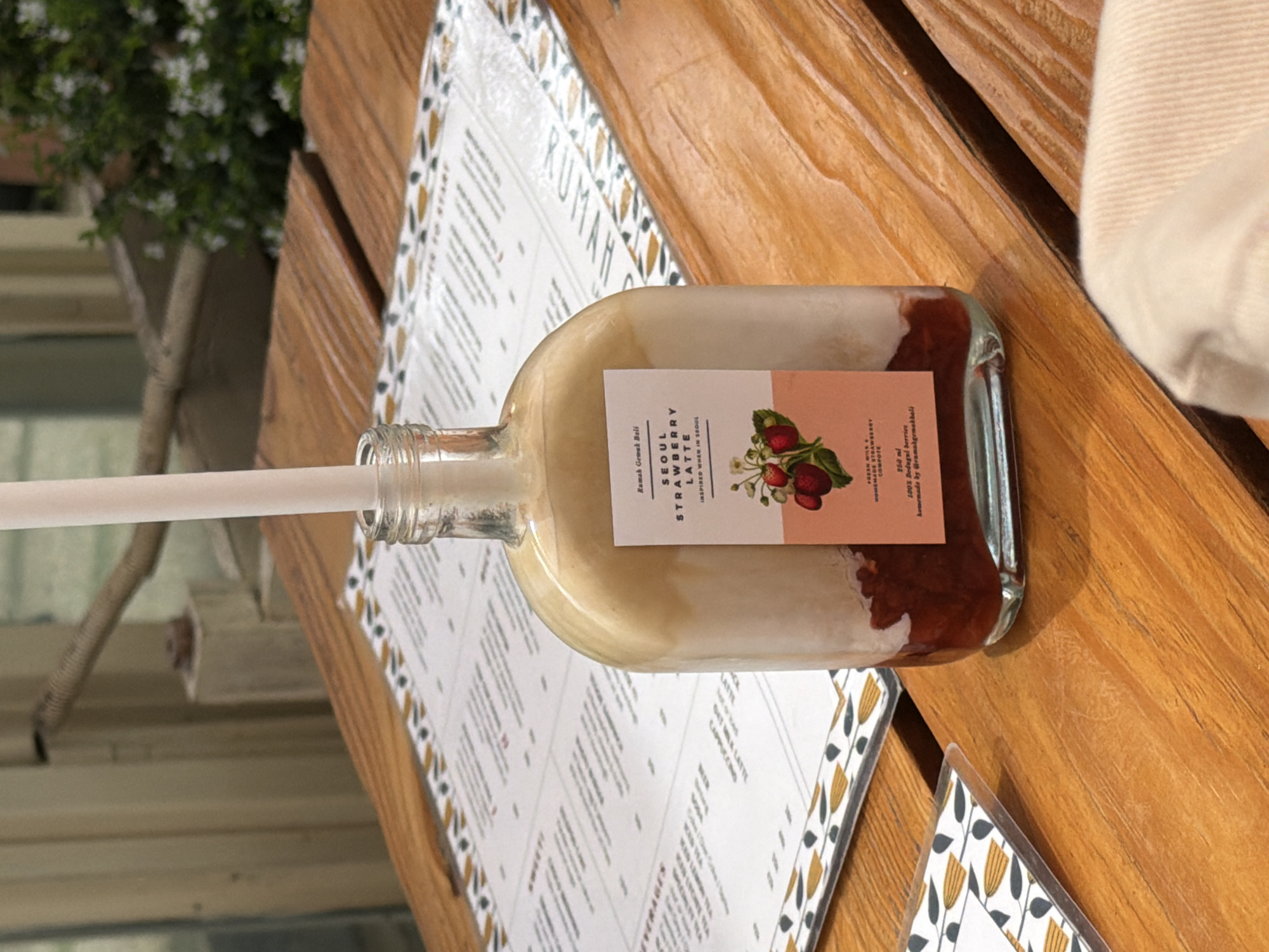 The image captures a strawberry latte drink in a glass bottle on a table at Rumah Gemak Bali, a restaurant in Candikuning, Indonesia. The table has a printed menu with a floral design. A white straw sticks out of the bottle. A portion of a person's white shirt can be seen in the top-right corner. The table is made of light brown wood. A plant is visible in the background near a window.
