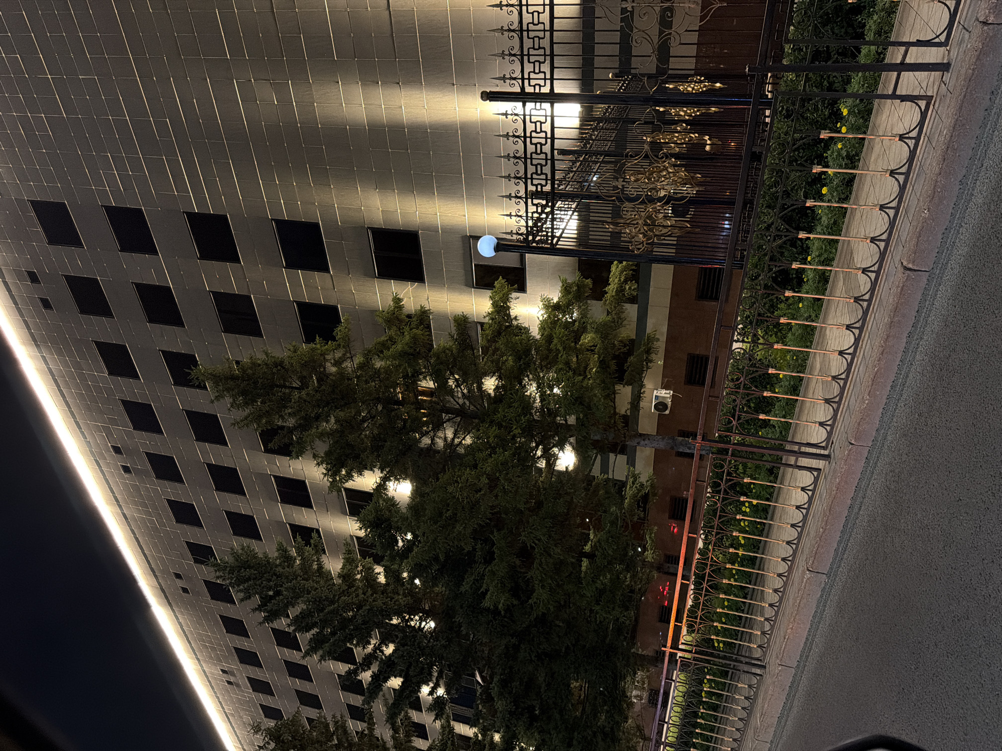 This is a nighttime, eye-level, outdoor shot of a modern, multi-story building with a metallic facade, located in Astana, Kazakhstan. The building is illuminated by a strip of warm white lights along its top edge and by vertical spotlights that cast shadows on its textured surface. The facade features a grid of dark, rectangular windows. In front of the building, to the left, are several tall, dense evergreen trees. A decorative black metal fence with ornate gold elements runs along the foreground, separating a sidewalk from the street. The street is paved and appears wet, reflecting the ambient light. Yellow flowers are visible in a garden bed behind the fence. A spherical blue light fixture is mounted on a pole near the fence. There are no people visible in the image.