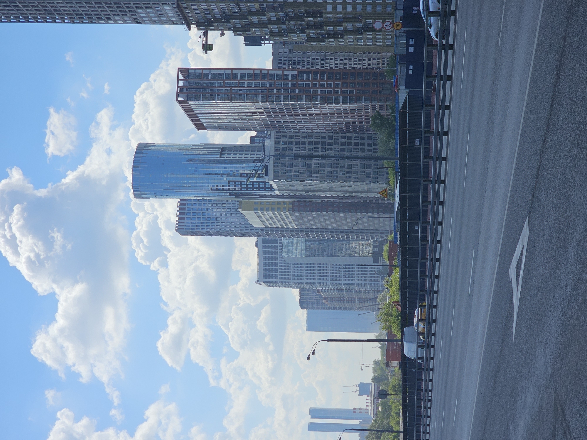 This media file captures a vibrant urban landscape under a bright, cloudy sky. The scene is dominated by numerous high-rise buildings, indicative of a modern metropolitan area, specifically Moscow, Russia.
In the foreground, a multi-lane asphalt road spans the lower portion of the frame. White dashed lines delineate the lanes, and a clear white arrow pointing forward is painted on one of the lanes. A dark metal barrier runs along the left edge of the road, separating it from the city structures.
The skyline is populated by a variety of contemporary high-rise buildings. Some boast sleek, reflective glass facades, while others display a mix of materials with numerous windows. A prominent, curved or cylindrical building with a highly reflective surface stands out in the mid-ground. Several construction cranes are visible among the buildings, indicating ongoing development.
Vehicles are scattered on or near the road, including a white van, a white car, and a partially visible yellow vehicle, suggesting typical urban traffic. Multi-armed streetlights extend over the roadway. Green foliage from trees is visible, interspersed among the buildings and lining parts of the road.
The sky is a clear blue, adorned with large, fluffy white cumulus clouds, indicating fair weather. The bright and even illumination suggests it is daytime, likely late morning or early afternoon. Visible text includes a circular yellow speed limit sign displaying "40" in black, and a triangular yellow warning sign. The overall impression is one of a dynamic and expanding city with modern infrastructure and architecture.