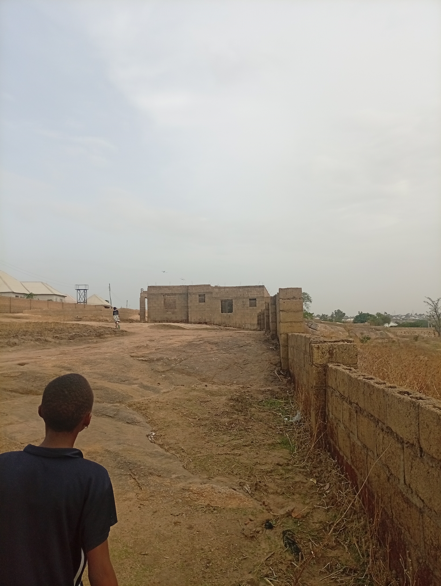 The image depicts an outdoor scene, likely a construction site or undeveloped area in Jos, Nigeria, given the presence of partially built structures and the environment. In the foreground, a person wearing a dark blue shirt is facing away from the camera, with the back of their head visible. To the right, a brick wall stretches into the distance, running along a dirt path. In the midground, a building under construction stands prominently, made of what appears to be cement blocks. The surrounding area consists of barren land, rocks, and sparse vegetation. In the background, rooftops of other buildings are visible, along with a water tower-like structure. Another person is walking in the distance. The sky is overcast.