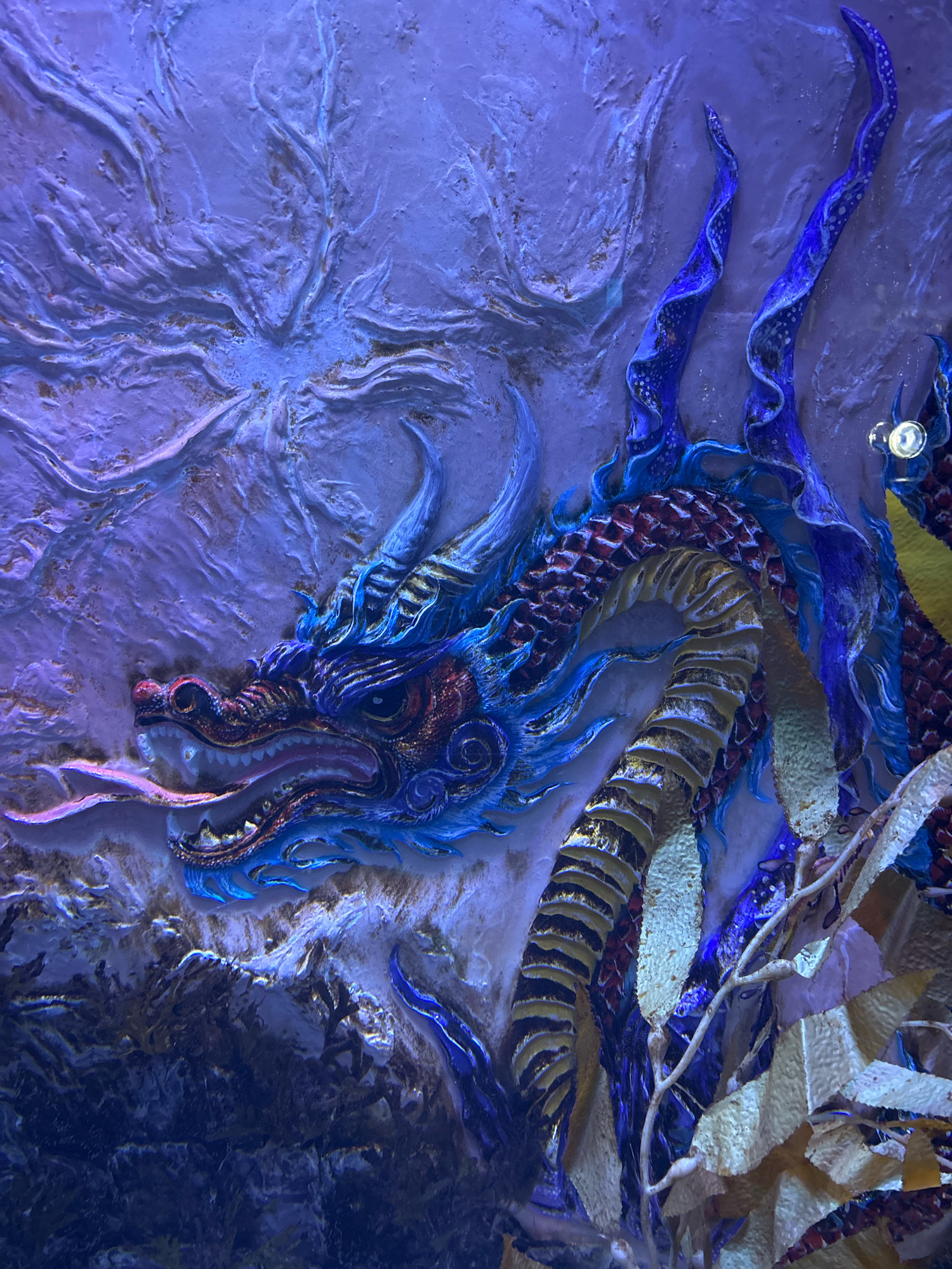 The image depicts a close-up of a detailed, three-dimensional relief sculpture of a dragon, illuminated by vibrant blue and purple lighting. The dragon's head is prominent on the left side of the frame, with its mouth open, revealing sharp teeth and a long, pink tongue. Its scales are intricately rendered, with shades of blue, red, and gold. The dragon's body coils towards the right, showing segmented sections and flowing blue elements resembling flames or water. The background appears to be a textured wall, possibly part of an aquarium or an artistic installation, with a swirling, almost cosmic pattern in shades of purple and white. In the lower right portion of the image, some translucent, plant-like elements, possibly seaweed or artificial decorations, are visible, adding to the aquatic or mystical theme. A small, bright light fixture is visible on the upper right, contributing to the overall dramatic lighting. This artwork is located in the United Arab Emirates' National Aquarium, located in the Qasr Al Hosn area of Abu Dhabi.
