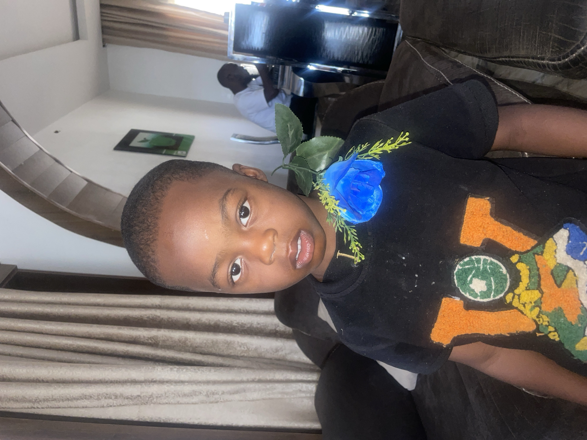 A young Black boy with closely cropped hair is shown indoors, wearing a black T-shirt with a colorful design. A bright blue artificial flower is pinned to the shirt near his shoulder. The background is blurred but shows a man in a white shirt sitting at a dark-colored desk and equipment that appear to be in a salon, and a staircase with beige striped curtains. The location is indicated as Jos, Nigeria.