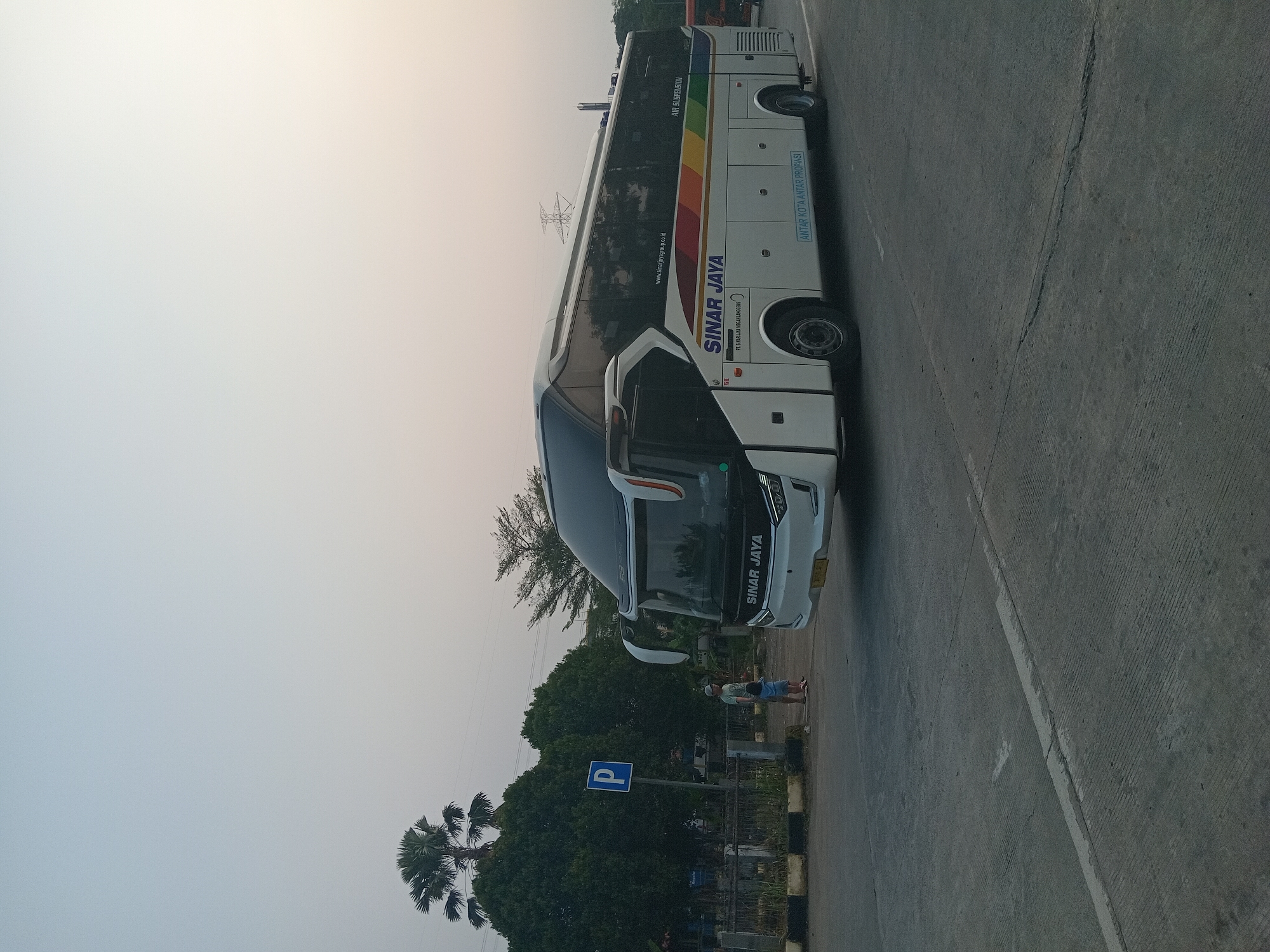 This image captures a bus labeled "SINAR JAYA" parked on a paved road, possibly in a rest area or parking lot. The bus is white with colorful stripes along its side. A "P" sign, indicating parking, is visible in the background, near a line of trees. Two individuals, an adult and a child, are standing near the sign, facing the bus. The sky is overcast, suggesting either early morning or late afternoon. The overall setting appears to be an outdoor urban environment.