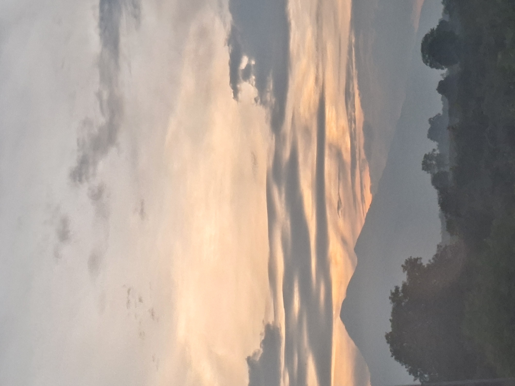 This is a vertical, full-frame photograph of a hazy mountain range in Keureuweungkrueng, Indonesia, during what appears to be either sunrise or sunset. The sky is filled with a gradient of soft peach and gray clouds, with distinct horizontal cloud bands that create a layered effect. Sunlight filters through some of these clouds, casting subtle beams. In the foreground, silhouetted against the sky, is a dense band of trees and foliage, with a prominent, large tree in the lower left. Behind the trees, a large, dark mountain with a rounded peak dominates the middle ground. The overall atmosphere is serene and slightly obscured by atmospheric haze. No people are visible in the image.