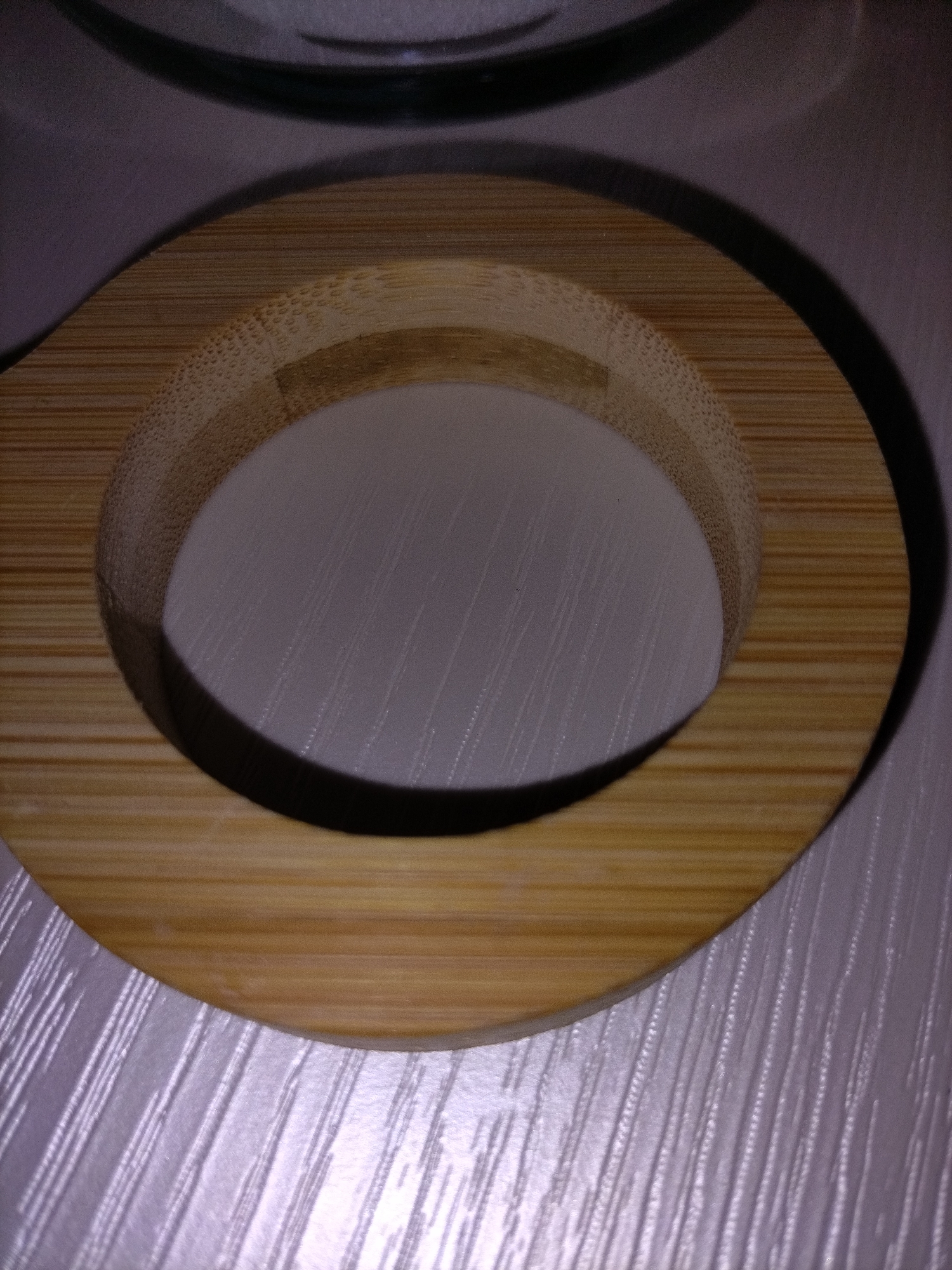 The image shows a close-up, overhead view of a circular bamboo object, possibly a coaster or a lid, resting on a textured white surface. The bamboo has a natural wood grain pattern with visible lines and subtle color variations. The object is casting a shadow on the surface below, indicating a light source from above and to the side. The white surface appears to have a raised, parallel-line pattern, creating a ribbed or corrugated texture. The overall lighting is dim, with some areas brighter than others due to the light source. There are no people or animals visible in the image.