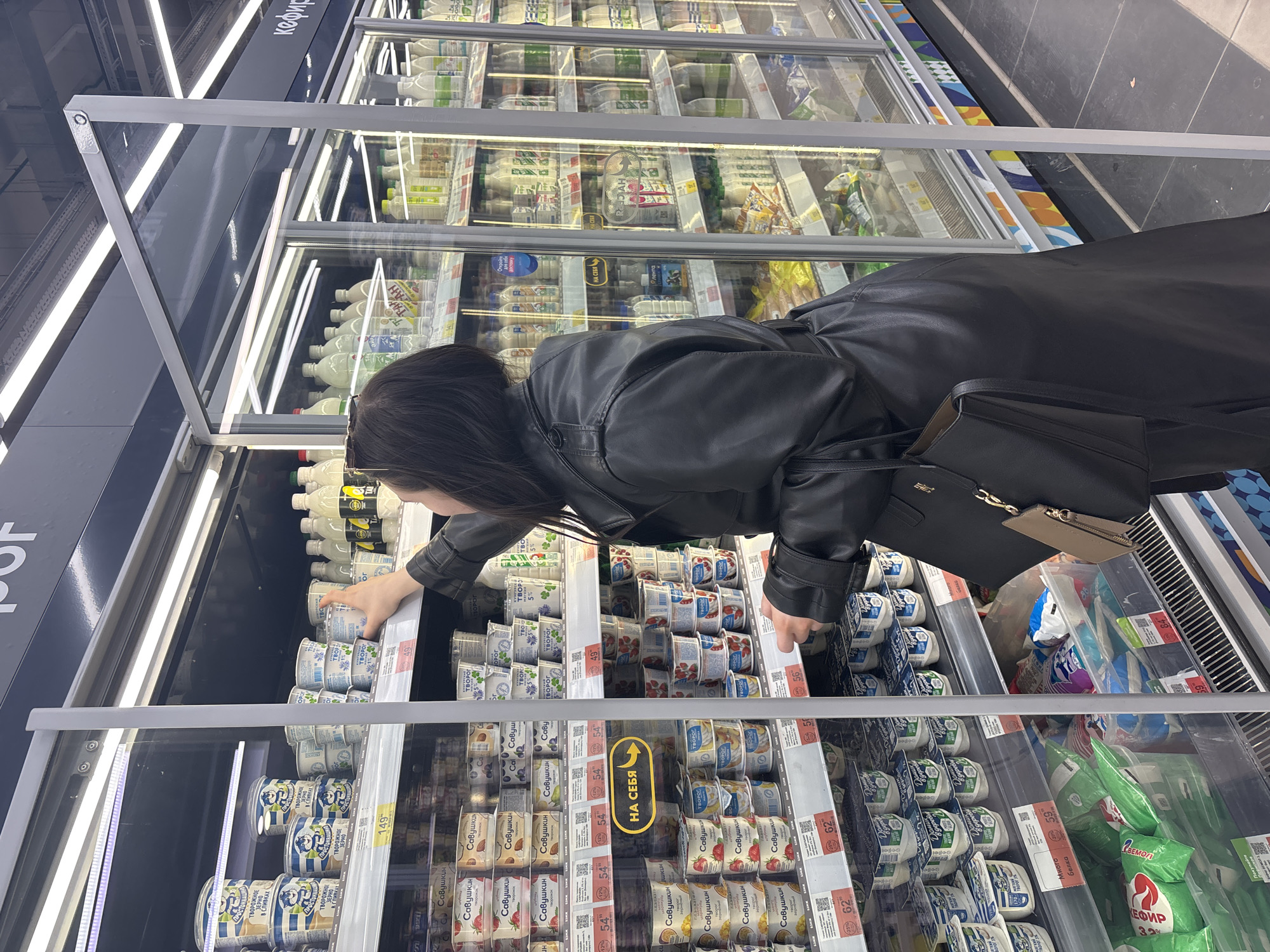 A woman with short dark hair is reaching into a refrigerated dairy case in a supermarket. She is wearing a black leather jacket and has a black purse over her shoulder. She is selecting dairy products, specifically small containers of cottage cheese and yogurt. The dairy products in the case are labeled in Russian. In the foreground is a sign in Russian indicating how to open the door. The products on the shelf include "Savushkin" brand cottage cheese with various fruit flavors. The location is likely a supermarket in Perm, Russia, as the products are labeled in Russian and the supermarket chain "Lenta" is identified.