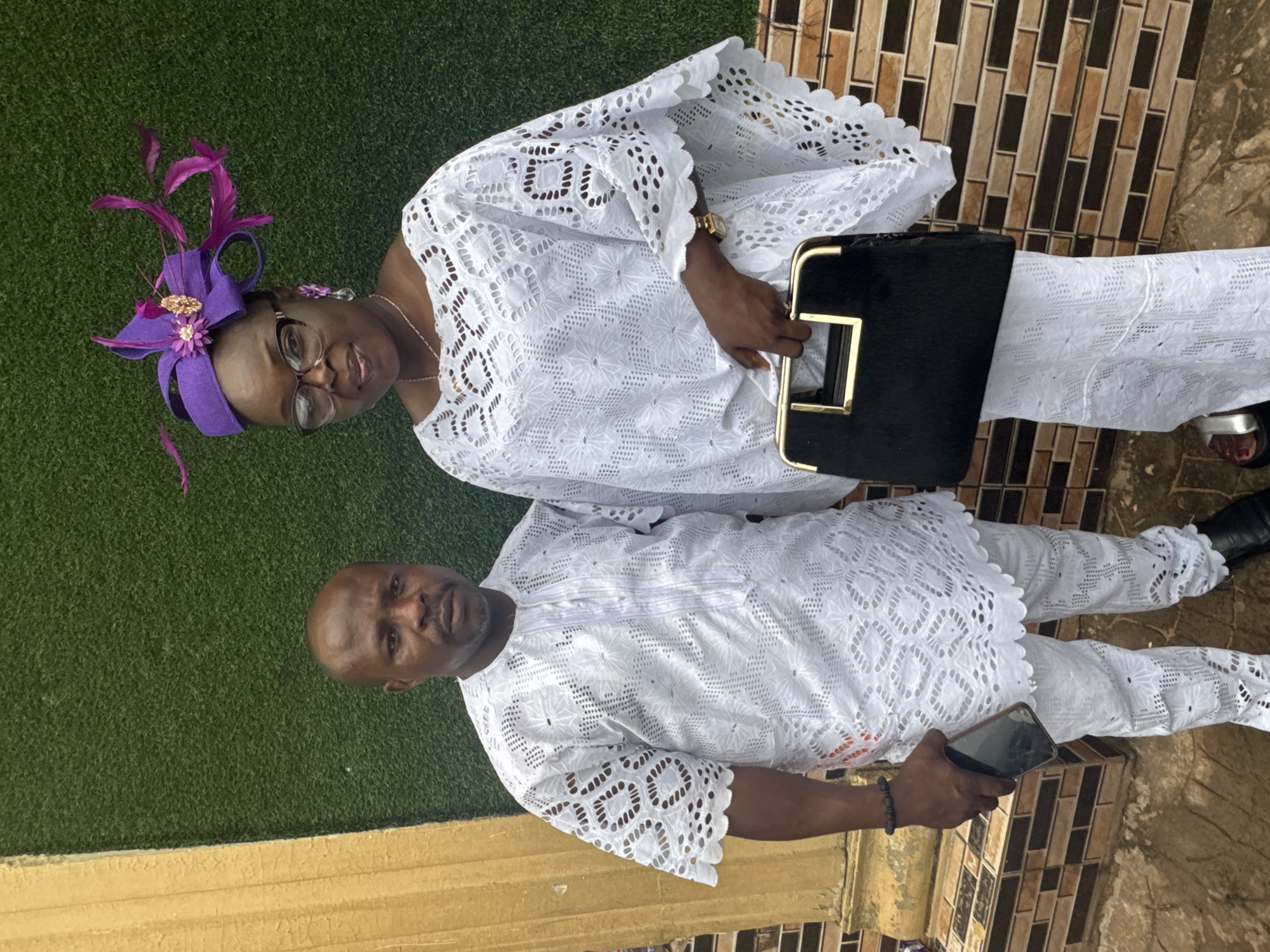 The image shows two individuals standing outdoors, a woman and a man, both dressed in matching white lace outfits. The woman is wearing a purple fascinator with feathers and eyeglasses, and is holding a black handbag with gold accents. The man is holding a smartphone. They are standing on a surface of artificial turf next to a wall with tan stone and another area with tile. The location is in Nkpor, Nigeria.