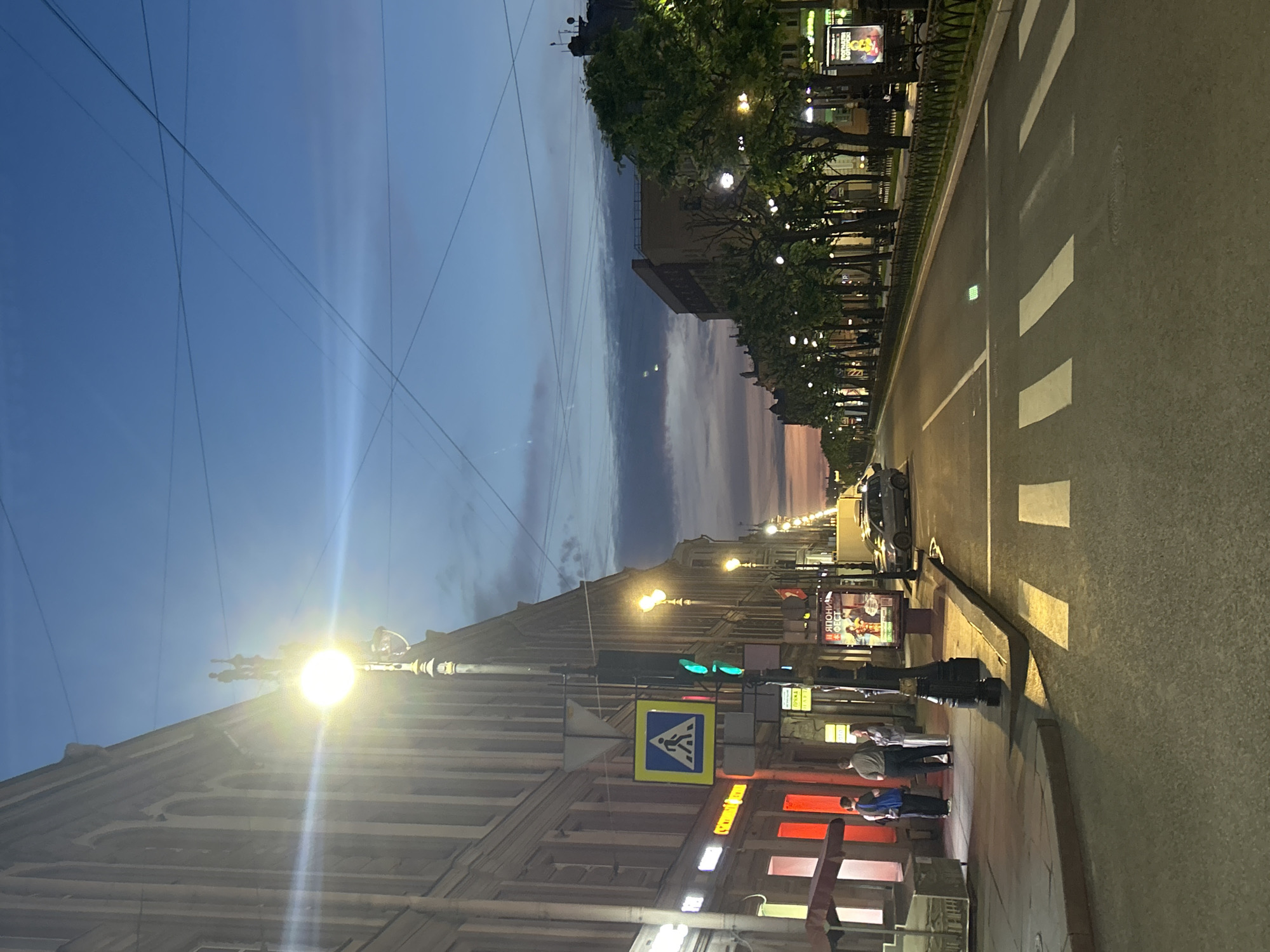 The image depicts a street scene at dusk or dawn. On the left, a building with arched windows and ornate detailing is visible, illuminated by streetlights. A pedestrian crossing sign is mounted on a pole in front of the building, alongside a traffic light showing green. Several people stand on the sidewalk near the building.
The street itself is paved, with white pedestrian crossing markings clearly visible in the foreground. To the right of the street, a row of trees with green foliage lines the sidewalk, interspersed with streetlights. Beyond the trees, more buildings and structures are visible, some with lit windows. The sky above is a gradient of blue and pink, with visible overhead wires. A car is parked on the right side of the street. A poster advertising a "Japanese Fest" is displayed on a billboard. The overall impression is of a city street transitioning from day to night or vice versa.