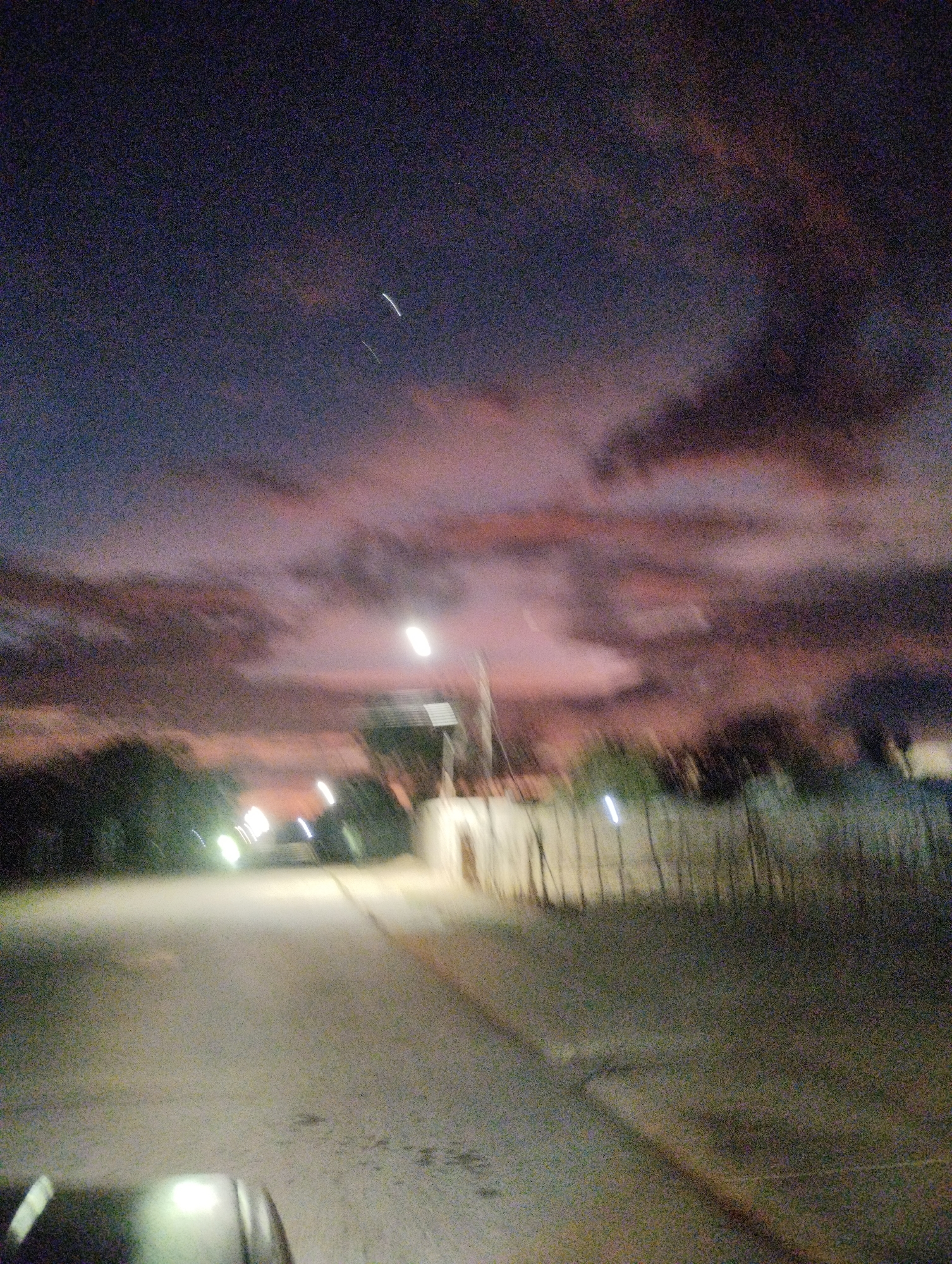 The image shows a street scene at dusk or dawn in La Guardia, Venezuela. The sky is a mix of dark blues and purples with streaks of pink and orange from the setting or rising sun, illuminating wispy clouds. Two faint, bright lines, possibly representing aircraft or satellites, are visible in the upper sky. The foreground features a paved road with some debris or markings. To the right of the road, there is a fence made of vertical wooden posts, partially obscured by foliage. In the background, blurred lights suggest buildings and trees. The overall impression is one of a quiet, rural or semi-rural setting during twilight.