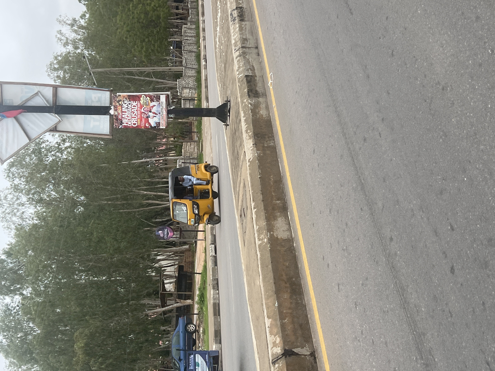 A yellow auto rickshaw with a driver is driving down a paved road in Jalingo, Nigeria. The rickshaw has a single passenger in the back. A blue sedan is parked on the side of the road. There are trees and a billboard in the background. The billboard shows a poster for a "Healing Deliverance Crusade" in Jalingo. The road is lined with a concrete barrier.