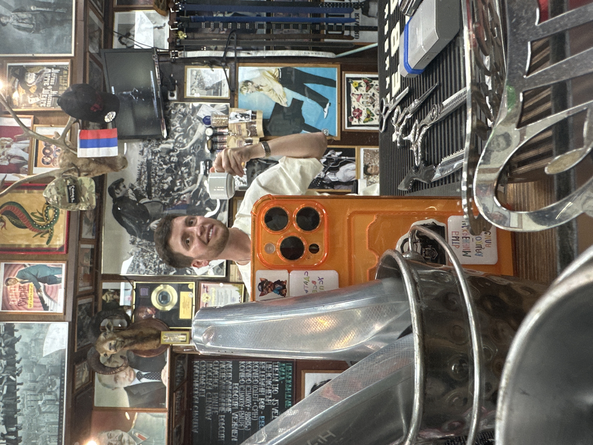 A man is taking a selfie in what appears to be a cluttered interior. The location is likely a barbershop in Moscow, Russia, as indicated by the Russian language on a menu-like board. The walls are covered with pictures, posters, and taxidermied animal heads. The man is holding an orange phone with a clear view of his face, indicating he is the subject of the selfie. Metal barber tools are visible in the foreground. The environment is densely decorated and suggests a vintage or retro aesthetic.