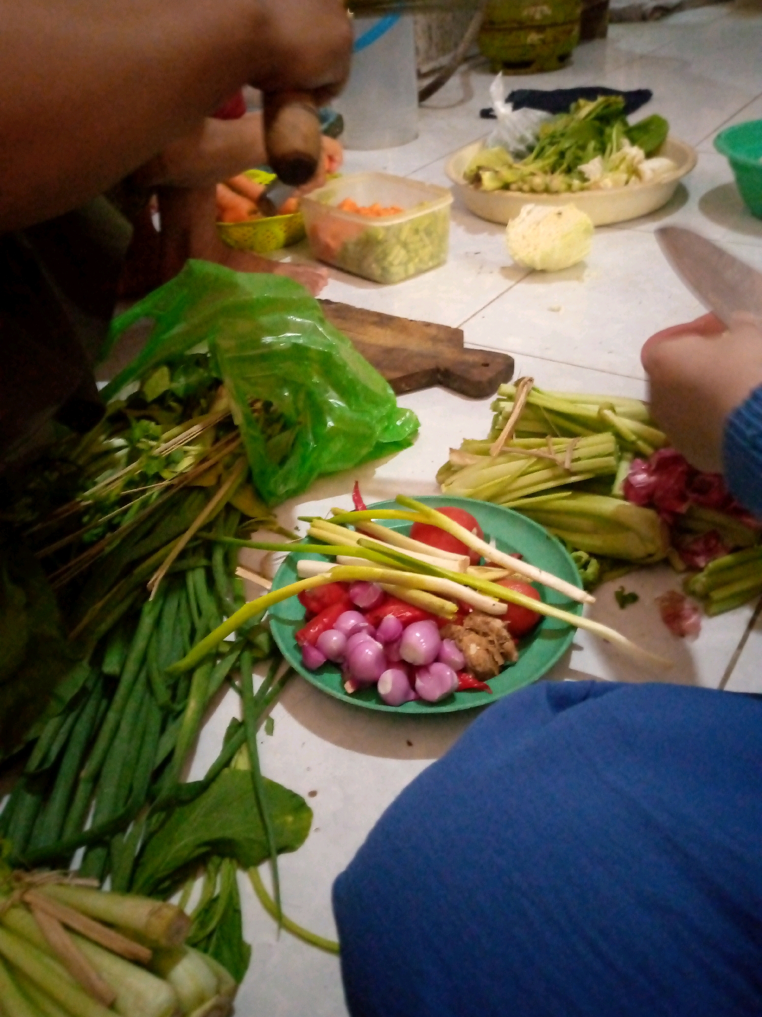 A low-angle, eye-level shot shows a person's hands preparing vegetables in a room in Sedati, Indonesia. The foreground features a blue cloth and a green plate piled with chopped tomatoes, shallots, and green onions. To the left, a bunch of long green vegetables, possibly scallions or leeks, lies on the floor next to a green plastic bag filled with more leafy greens. In the middle ground, a person's hands are visible, one holding a wooden handle that appears to be part of a mortar and pestle, possibly grinding spices. A wooden cutting board is also present. Further back, a bowl of chopped carrots and a larger bowl of mixed vegetables, including what looks like cauliflower and leafy greens, are on a light-colored tiled surface. The overall scene suggests a home kitchen environment where food preparation is underway.