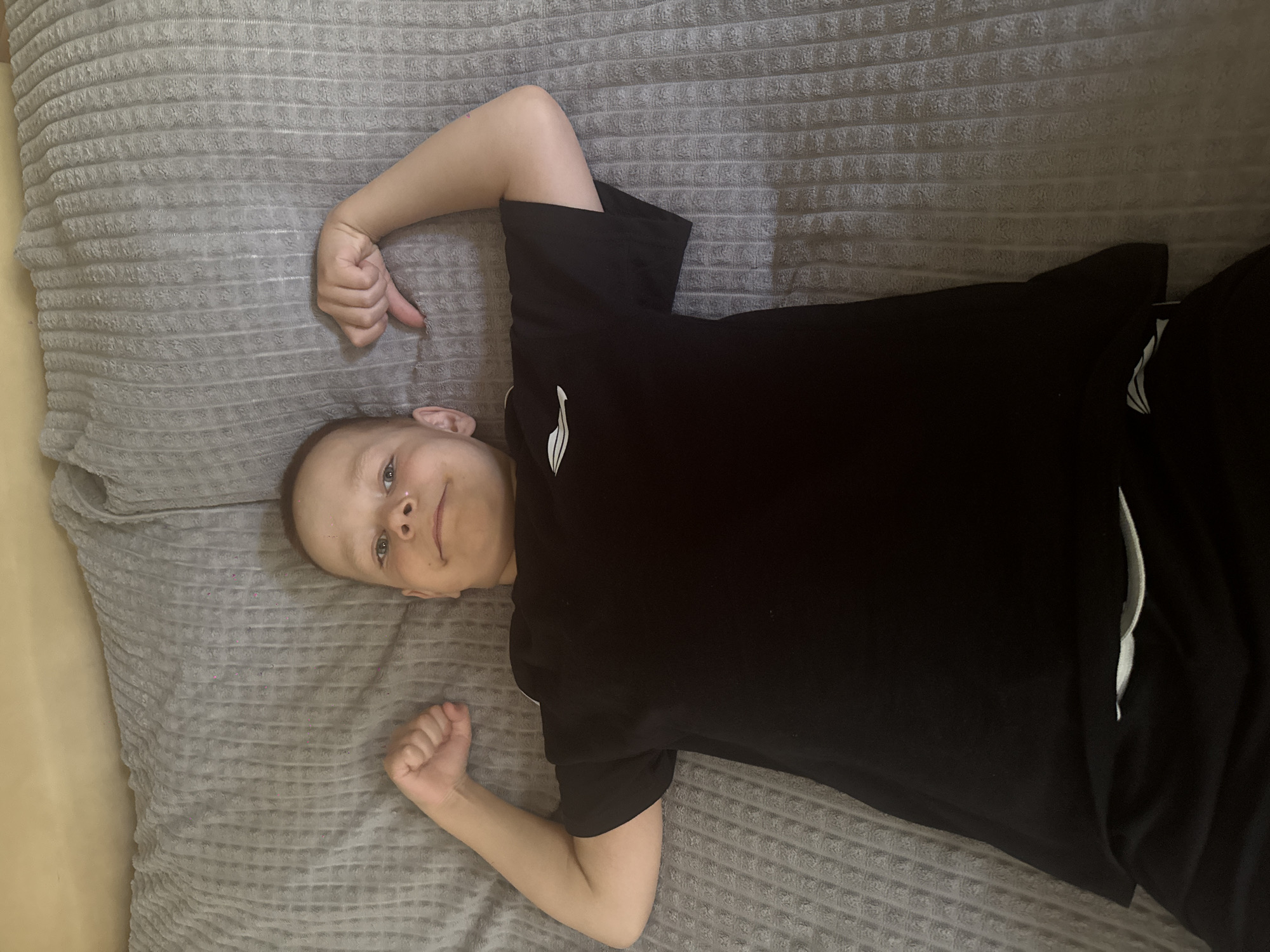 A young boy lies on a gray, textured surface, possibly a bed or couch. He wears a black t-shirt and black pants with a white stripe. The boy is positioned at an angle, with his head near the left side of the frame and his legs extending towards the right. His arms are bent, with his fists clenched near his head and waist. The location is Chaikovsky, Russia.