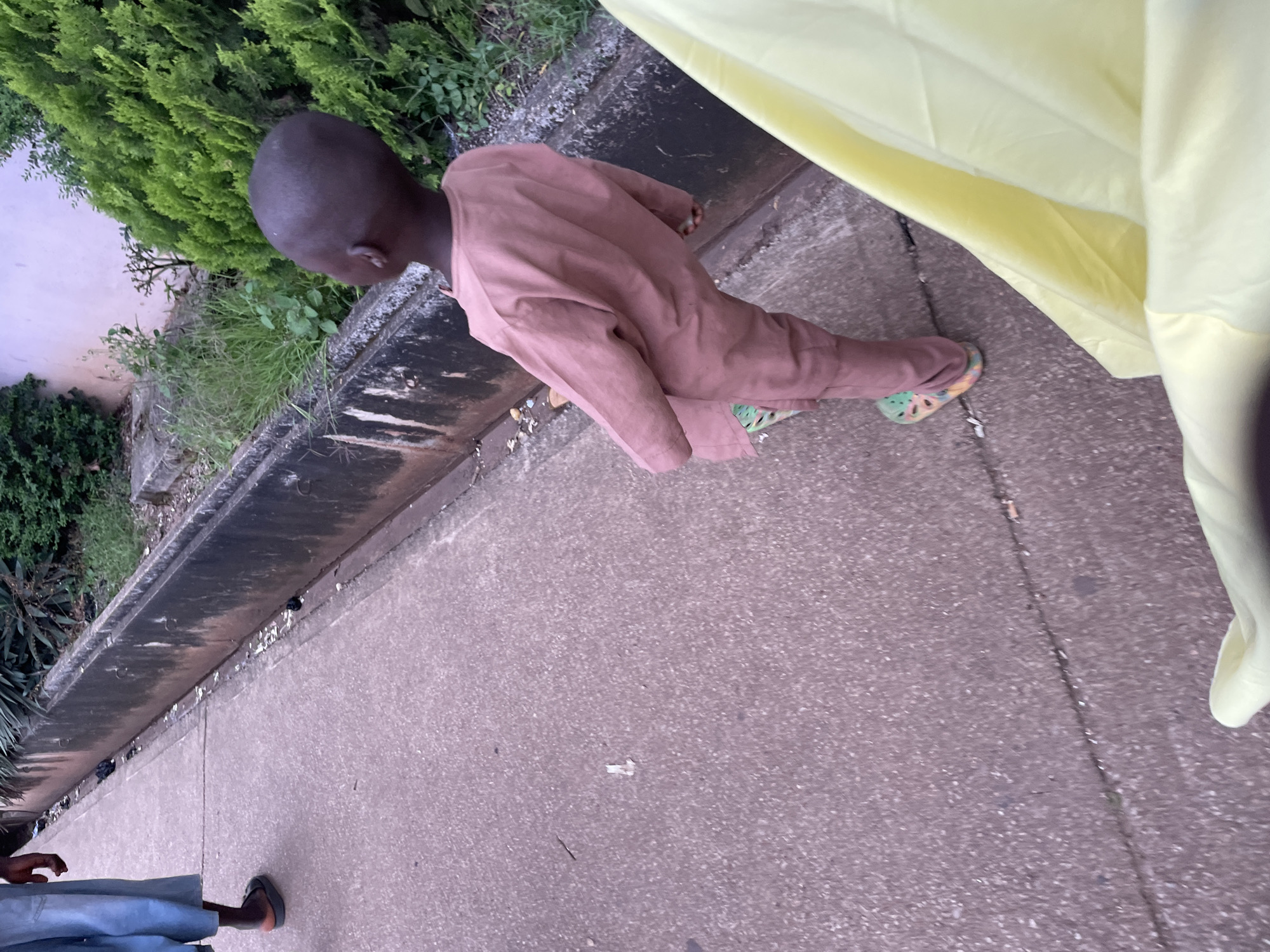 The image depicts a scene in Jos, Nigeria, with a young child wearing a light brown robe and multi-colored clogs standing on a concrete pathway. To the left, another person's legs are visible, wearing blue garments and sandals. The pathway runs alongside a concrete gutter filled with debris. Lush green bushes and trees are present in the background, with a pale pink wall behind them. The lighting suggests it is either early morning or late afternoon. The overall impression is of an outdoor, possibly residential or public area in a developing urban environment.