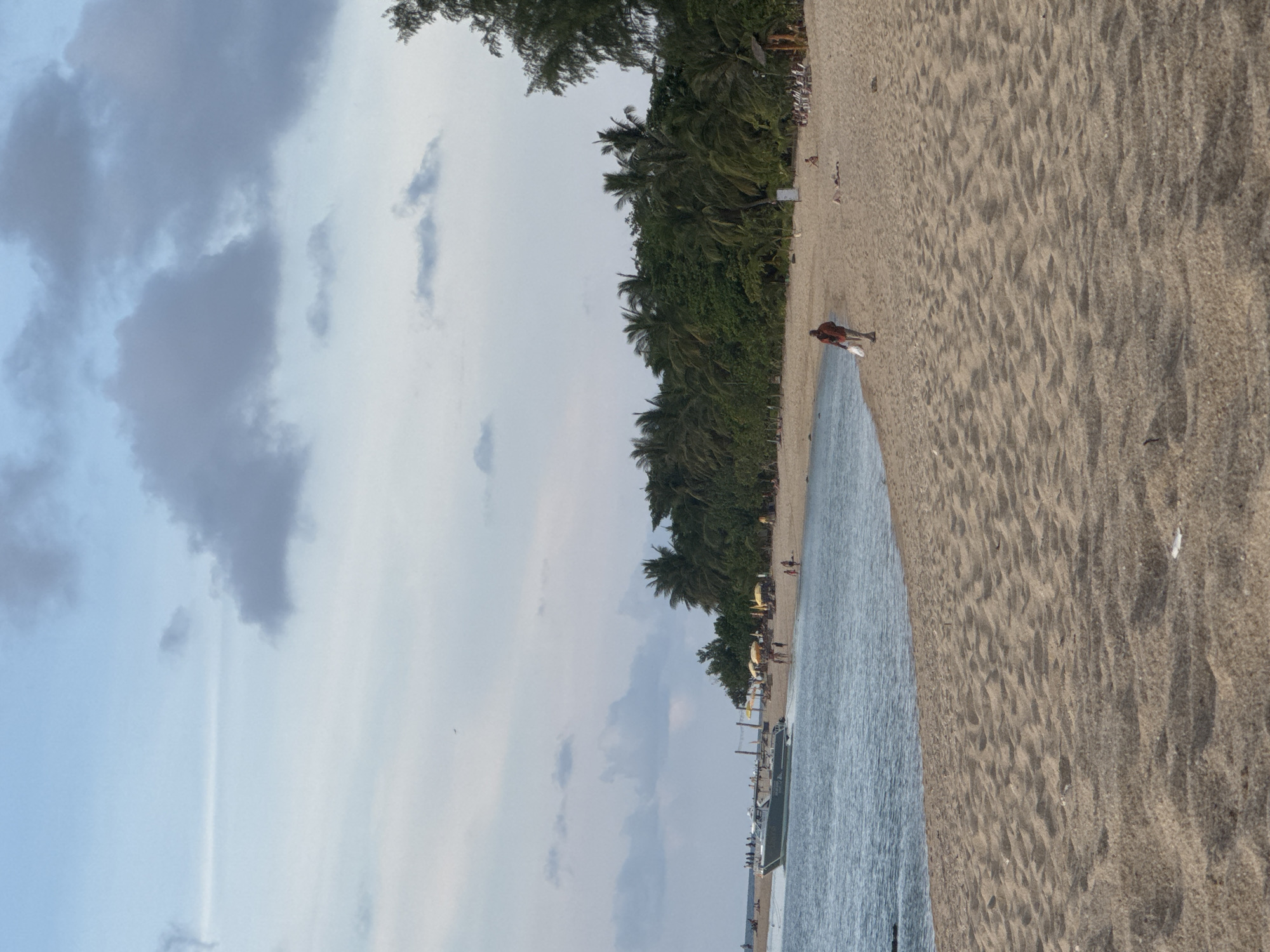 The image depicts a beach scene in Baan Wang Ta Kien, Thailand. A coastline of palm trees separates the sand from the sky. A person wearing a red shirt is walking along the beach with an object in their hands. The sand is tan, and the water is blue-grey. The sky is overcast with cloudy skies.