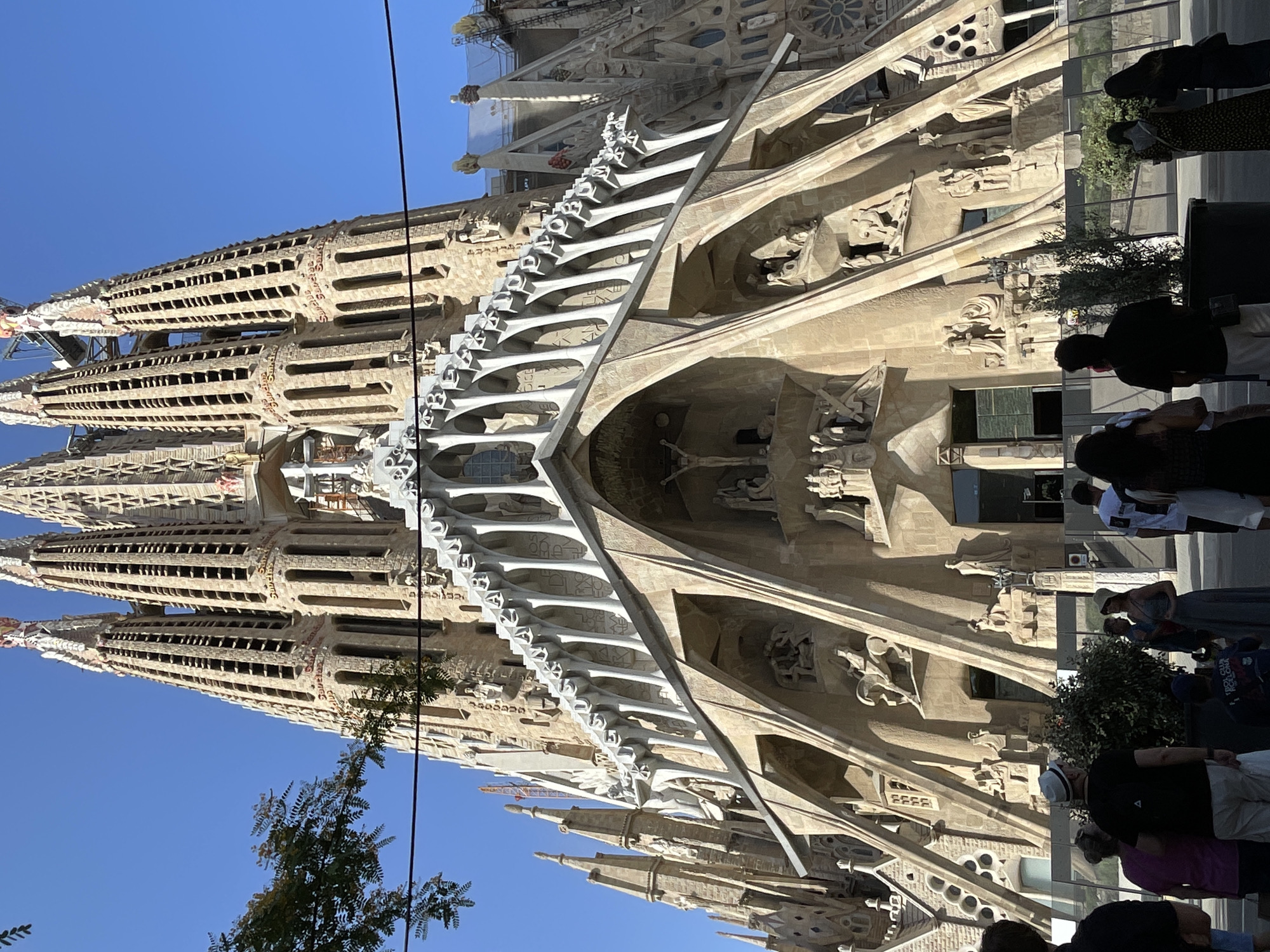 This is a low-angle, full-shot, outdoor photograph of the Sagrada Familia basilica in Barcelona, Spain, taken on a clear day. The basilica's facade is made of a light-colored stone and features intricate architectural details, including spires, arches, and numerous sculptures. Some of the sculptures depict religious scenes, such as a crucifixion. The facade is adorned with inscriptions. In the foreground, a group of about ten people are visible, standing and looking towards the basilica. They are of various ages and are dressed in casual clothing. Some are wearing hats. The ground in front of the basilica appears to be paved, and there is a large potted plant with green foliage. A black fence runs across the middle ground, separating the viewers from the construction area of the basilica. In the upper portion of the image, construction cranes are visible near the tops of the spires.