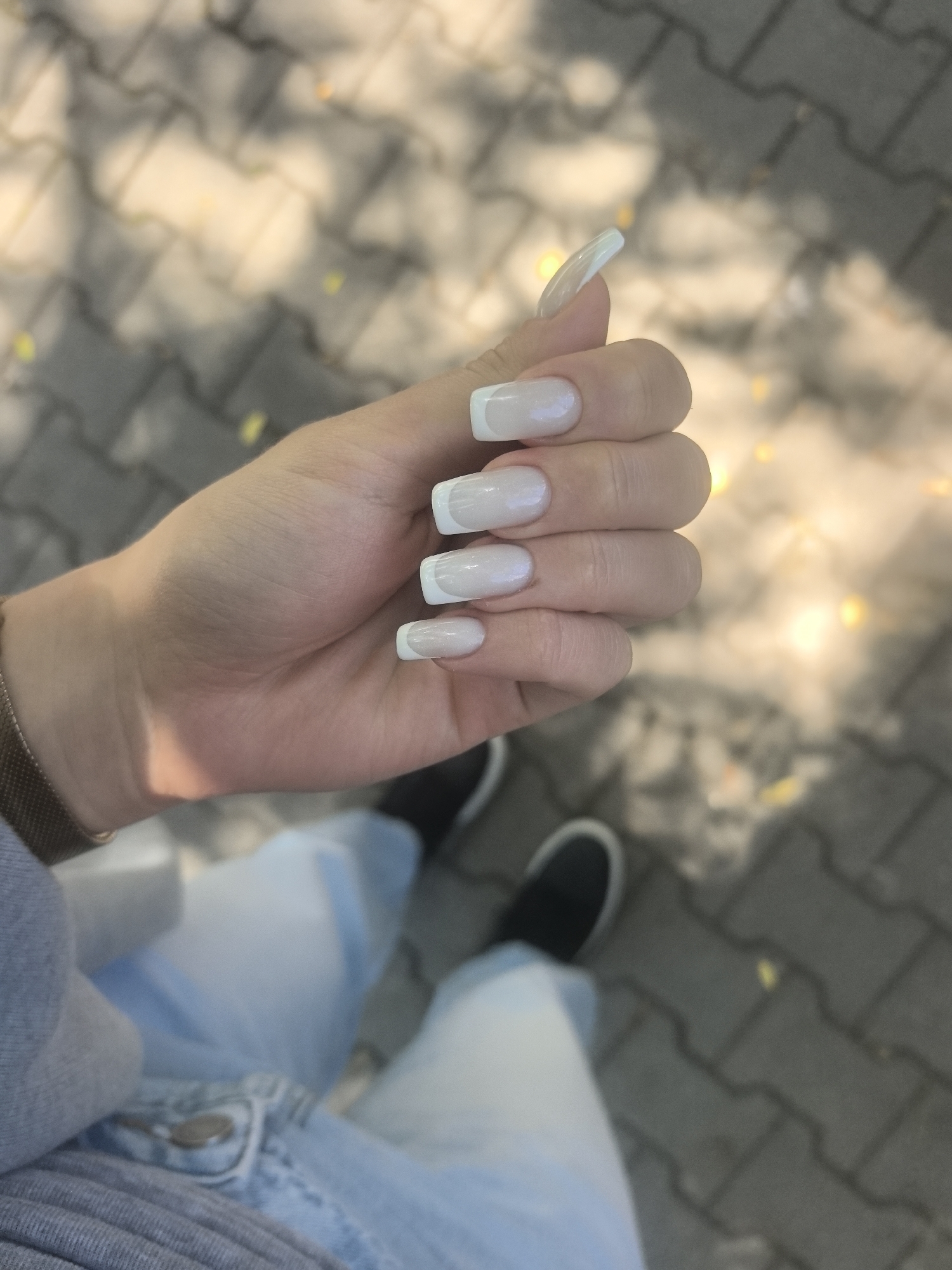 This image captures a close-up, first-person perspective looking down at a person's hands, lower body, and the ground in an outdoor setting in Stara Zagora, Bulgaria. The central subject is a light-skinned hand, held slightly angled, prominently displaying long, square-shaped nails with a classic French manicure. The nails feature a sheer, light pink or natural base with distinct white tips. The thumb nail is notably long and slightly curved.
On the wrist, a metallic mesh-strap watch is visible. Below the hand, portions of light blue jeans and a grey garment are discernible, along with black, low-profile shoes on the feet. The ground beneath is paved with grey interlocking concrete blocks, characteristic of sidewalks or pedestrian areas. The scene is brightly lit by natural sunlight, indicated by soft shadows cast on the hand and ground. The background is blurred, showing warm, yellowish-orange patches, possibly from sunlit foliage or reflections. There is no visible text in the image. The person appears to be posing to showcase their nails in a casual, daytime outdoor environment.