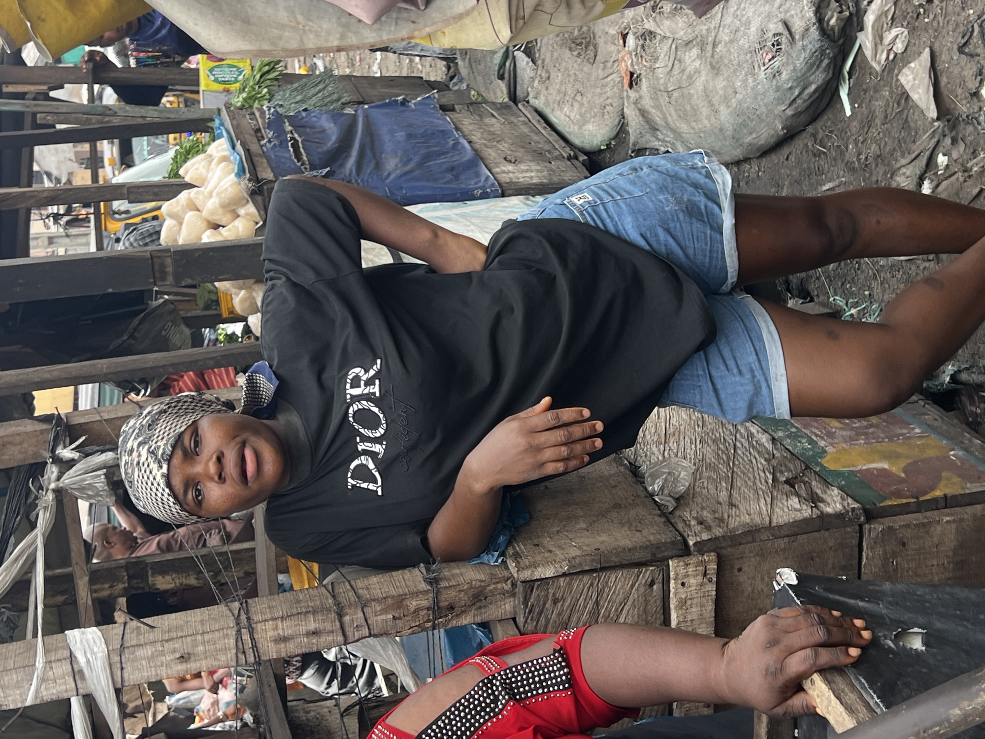 The image depicts a bustling outdoor market scene in Nkpor, Nigeria, captured during daytime. The central subject is a young woman, seen from the waist up, lying or reclining on a wooden market stall. She wears a black t-shirt with the word "DIOR" prominently displayed in white lettering across the chest, light blue denim shorts, and a patterned black and white headscarf. She looks directly at the camera with a neutral, slightly inquisitive expression. Her left arm is bent, resting on the wooden structure.
The market environment is characterized by makeshift wooden stalls and structures, giving a rustic appearance. Behind the woman, on an adjacent stall, are piles of light-colored, possibly processed food items such as garri or fufu, along with bundles of green leafy vegetables. Various sacks and blue tarpaulins, likely used for shelter or covering goods, are also visible. The ground below appears earthy, uneven, and somewhat cluttered with debris. In the immediate foreground, part of another person's arm and a striking red garment with silver studs are visible. Further in the background, indistinct market activity and parts of other structures or vehicles can be seen through the wooden framework. The overall impression is that of a lively, active local marketplace.