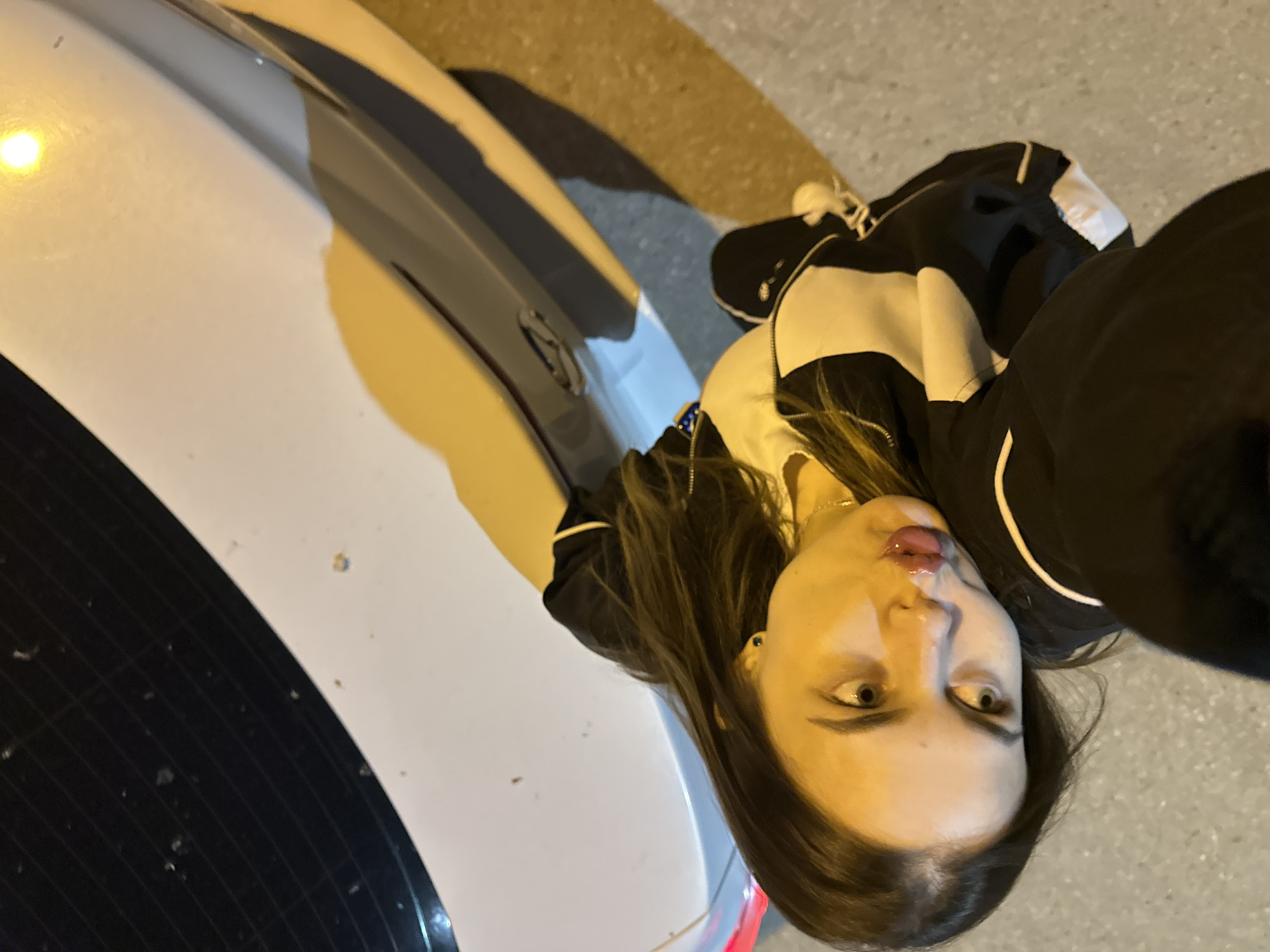 In the city of Perm, Russia, an image is captured from a high angle, looking down at a young woman with long brown hair. She is lying on her side, leaning against the trunk of a white car. The car's trunk lid is visible, with a black tinted rear window. The woman is wearing a black and white tracksuit. Her face is turned towards the camera, and her eyes are wide open. Her lips appear glossy, and she is resting her cheek on her hand, which is positioned near her chest. The ground beneath her is asphalt. The lighting suggests it is nighttime, with a yellow-orange glow illuminating the scene, likely from streetlights. A red taillight is visible on the far left of the car.