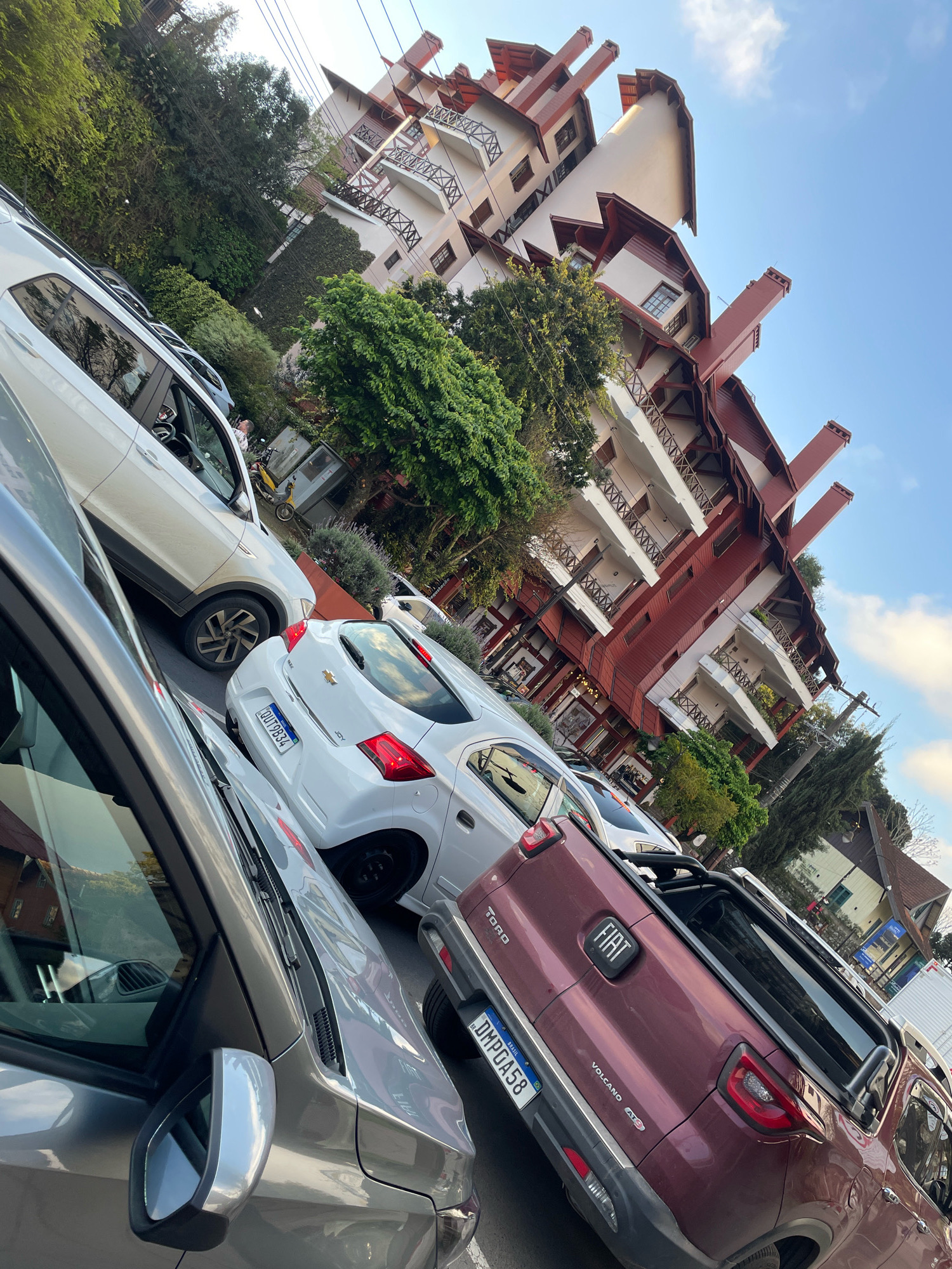 The image depicts a street scene in Gramado, Brazil, with a distinctive building in the background. The building is multi-story with a prominent red and white facade, featuring numerous balconies with wooden railings and several large red chimneys. The architecture suggests a European alpine influence, common in Gramado. In the foreground, several cars are parked along the street. A silver car is closest to the viewer, followed by a white Chevrolet hatchback and a maroon Fiat pickup truck. The license plates of the cars are visible. The street is paved, and there is vegetation, including trees and bushes, surrounding the area. The sky is partly cloudy and blue. The overall impression is a sunny day in a charming, well-maintained urban environment.