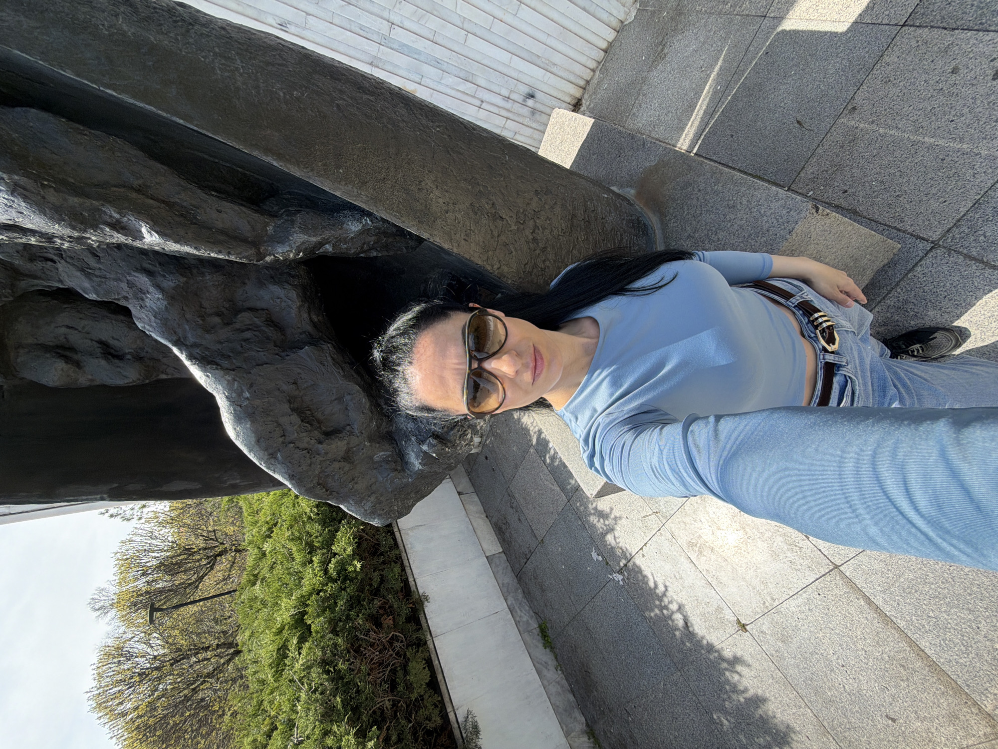 An outdoor selfie captures a woman posing next to a large, dark monument or statue in Pleven, Bulgaria, on a sunny day. The image is taken at an angle, with the woman's head positioned centrally towards the right side of the frame when corrected for orientation. She has long, dark hair and is wearing sunglasses, a light blue long-sleeved top that reveals a sliver of her midriff, light blue jeans, and a dark belt with a prominent gold buckle. Her expression is neutral to slightly smiling, looking directly into the camera.
A substantial, dark, textured statue or monument occupies the left side of the frame, suggesting a public square or historical site. The ground beneath her is paved with light and dark grey square tiles, showing distinct shadows cast by the monument and possibly trees, indicating bright daylight. In the background, particularly on the left, there is lush green foliage, including bushes and trees, with a lamppost visible among the branches. The clear skies and sharp shadows suggest pleasant, sunny weather. No visible text can be identified in the image.